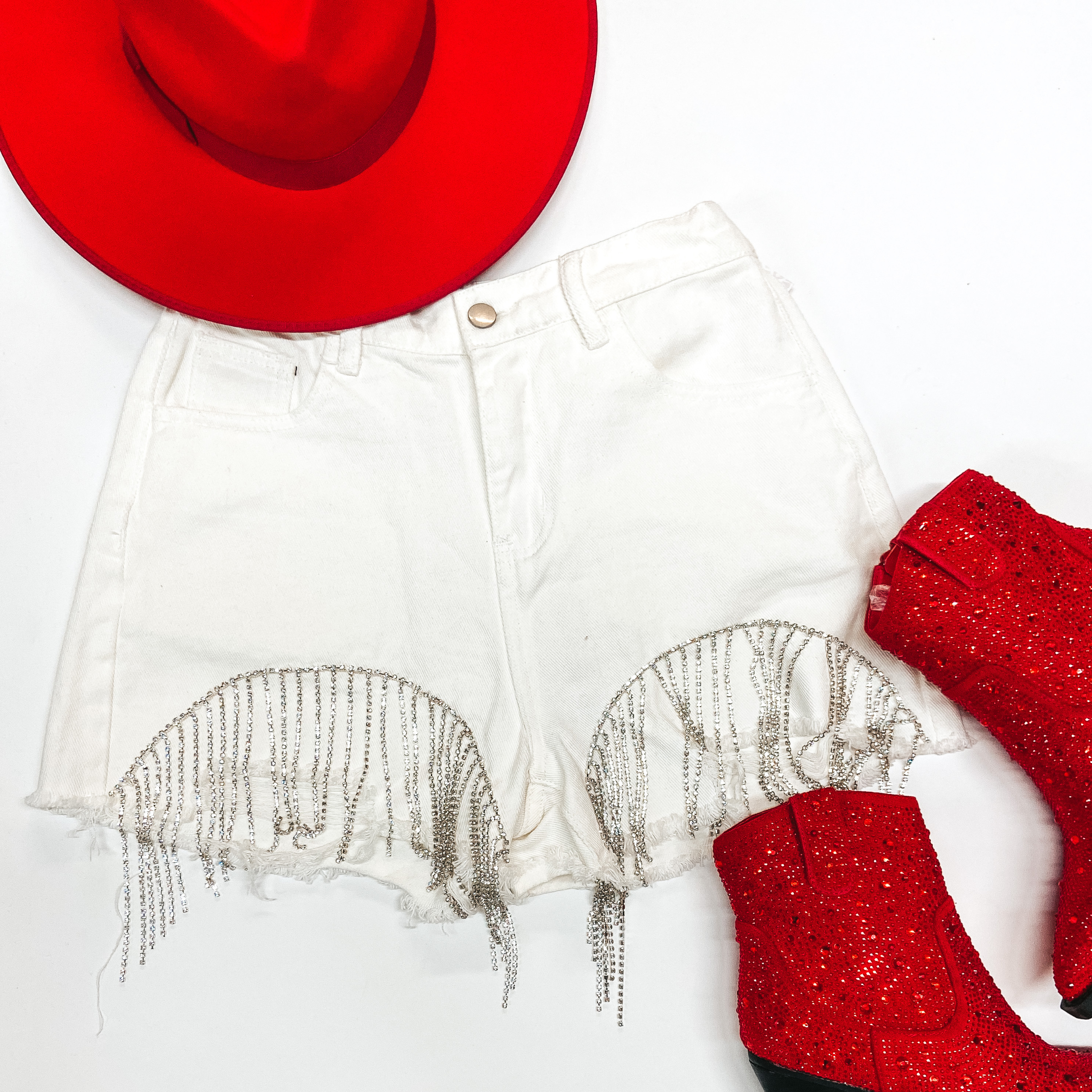 Ivory denim shorts with crystal fringe along the bottom. Paired with these shorts in the top left if a red Charlie 1 Horse hat and on the bottom right are red sparkly booties. Picture is on a white background. 