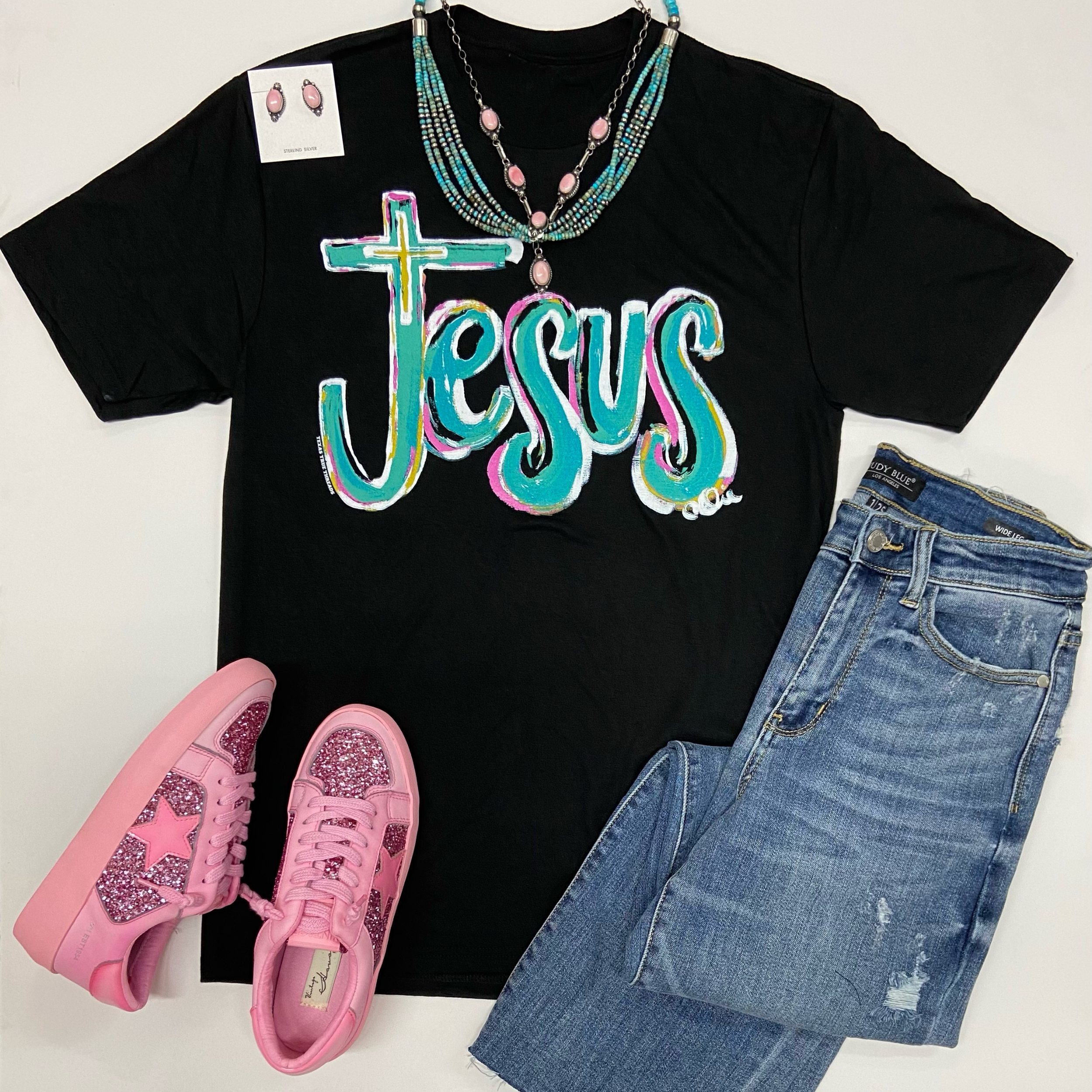 A black tee shirts with a crew neckline, short sleeves, and a graphic that says "Jesus" in turquoise writing. This tee is pictured on a white background with hot pink sneakers, Navajo jewelry, and light wash jeans.