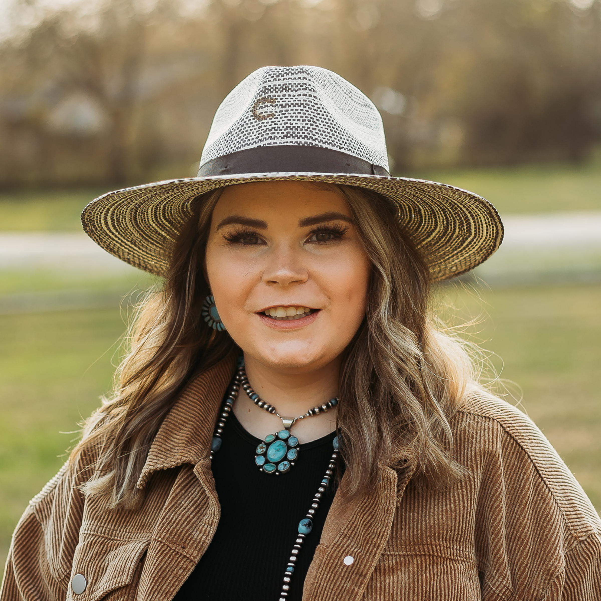 Black and White Straw Hat with Black Hat Band and Charlie 1 Horse Pin. Model has it paired with a brown jacket and turquoise jewelry. Pictured on wooded background