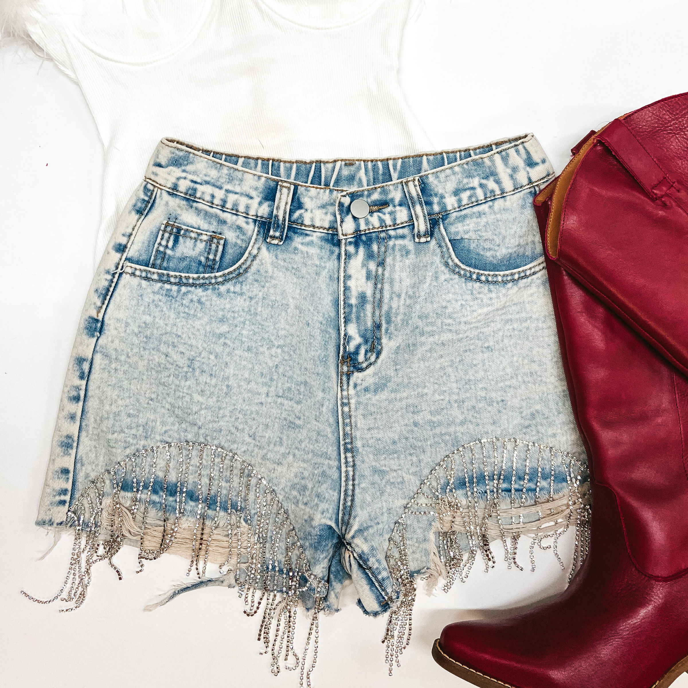 Light-wash denim shorts are centered in the middle of the picture with crystal fringe around the bottom. Red boots aew to the right of the shorts. All on a white background. 