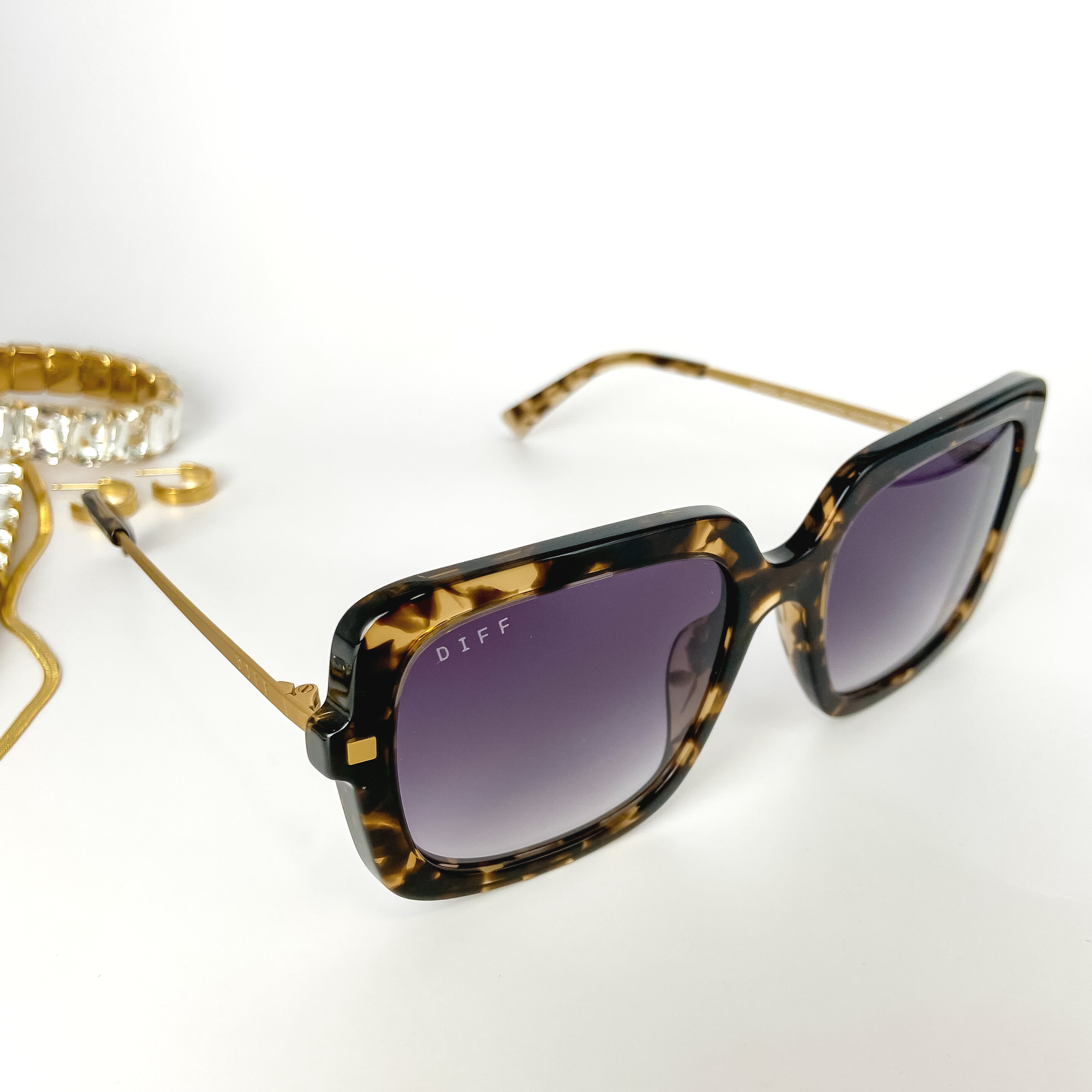 A pair of square tortoise-print sunglasses with gold tone trim and blue lenses. These sunglasses are pictured open on a white background with gold jewelry.