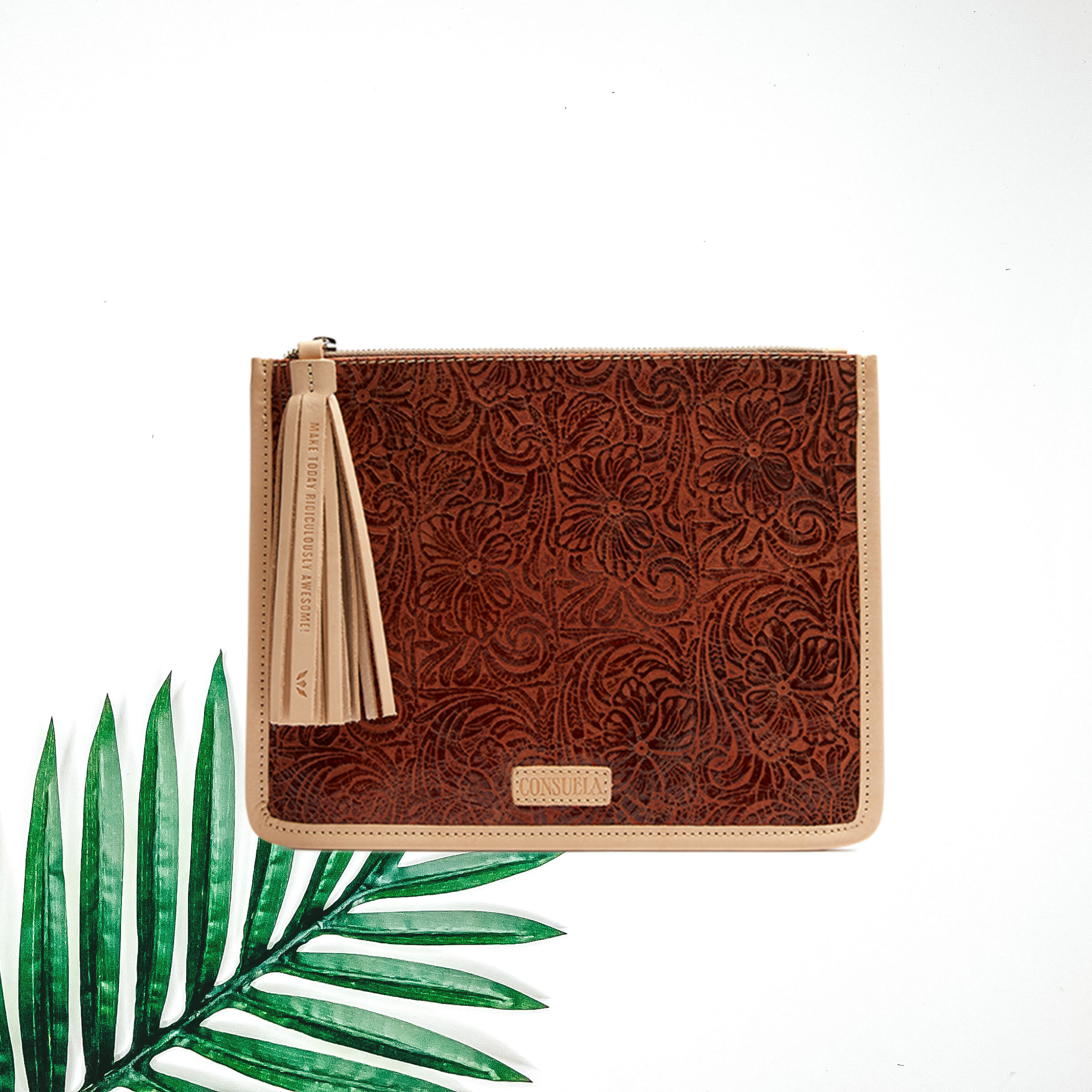 Centered in the picture is a pouch in a brown tooled pattern. To the left of the pouch is a palm leaf, all on a white background.