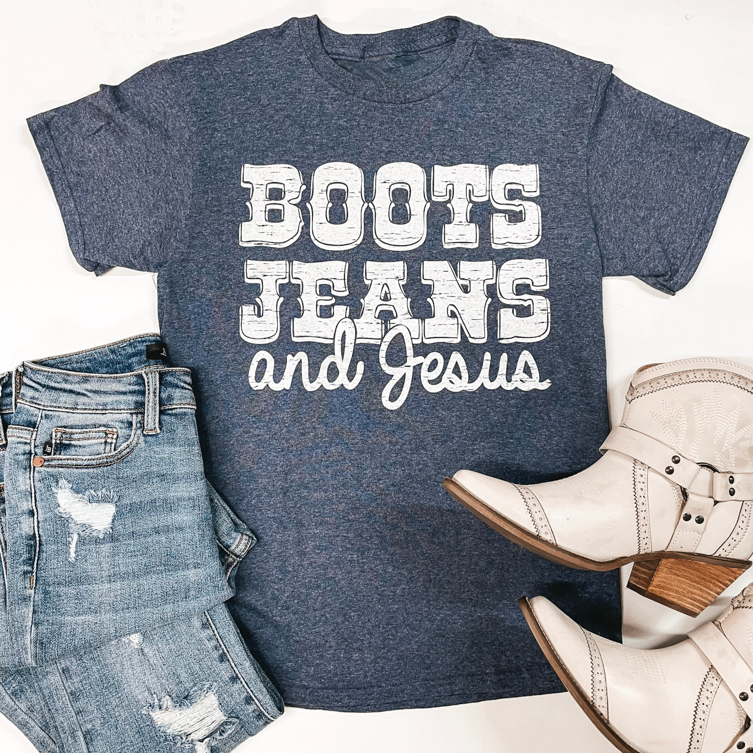 A light navy shirt is laid in the middle of the picture on a white background. In the center of the shirt is block letters stating: Boots Jeans and Jesus. On the bottom left of the picture is light-wash skinny jeans and on the bottom right is white boots. 