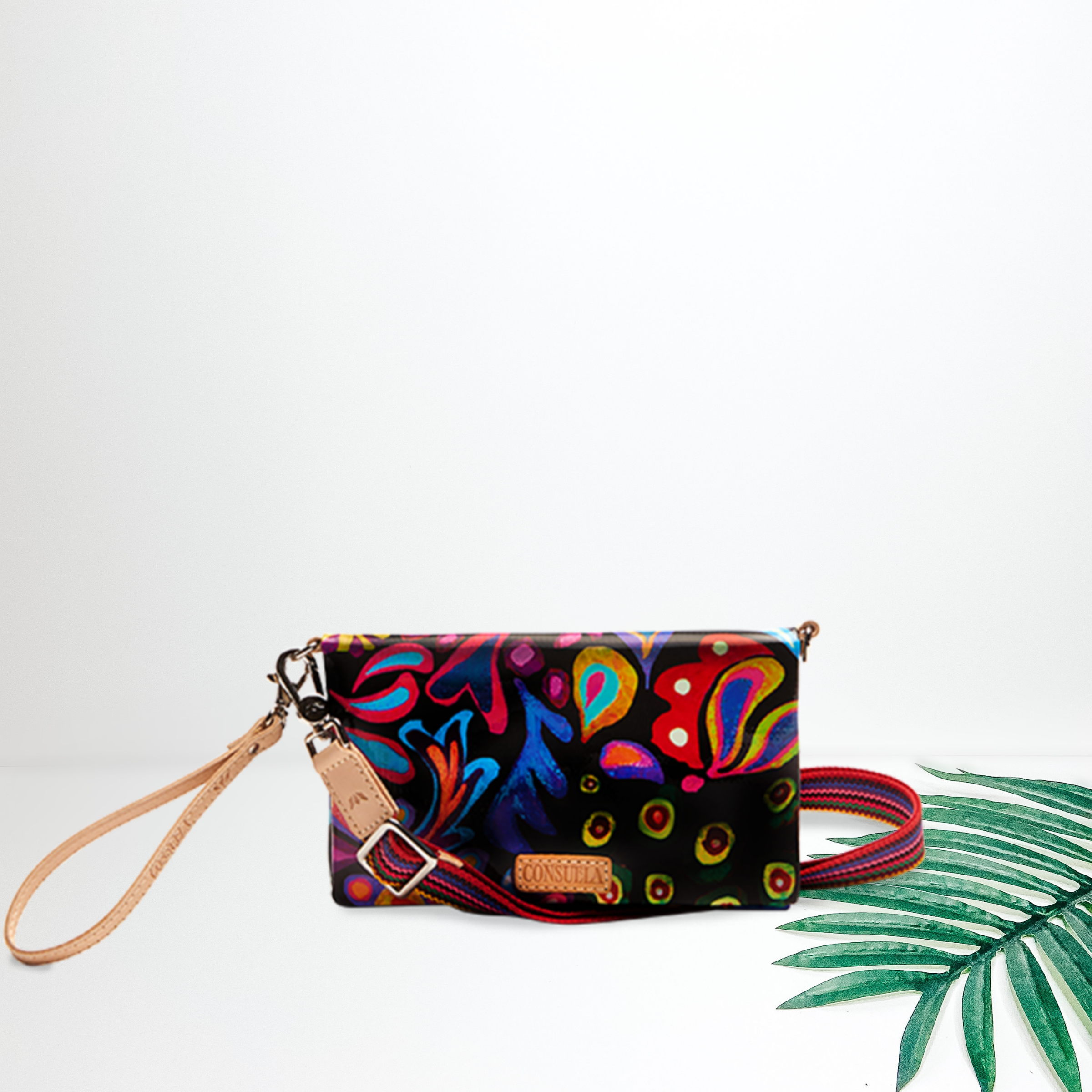 A black crossbody bag with a multicolor paisley print. To the right is a palm leaf, all on a white background