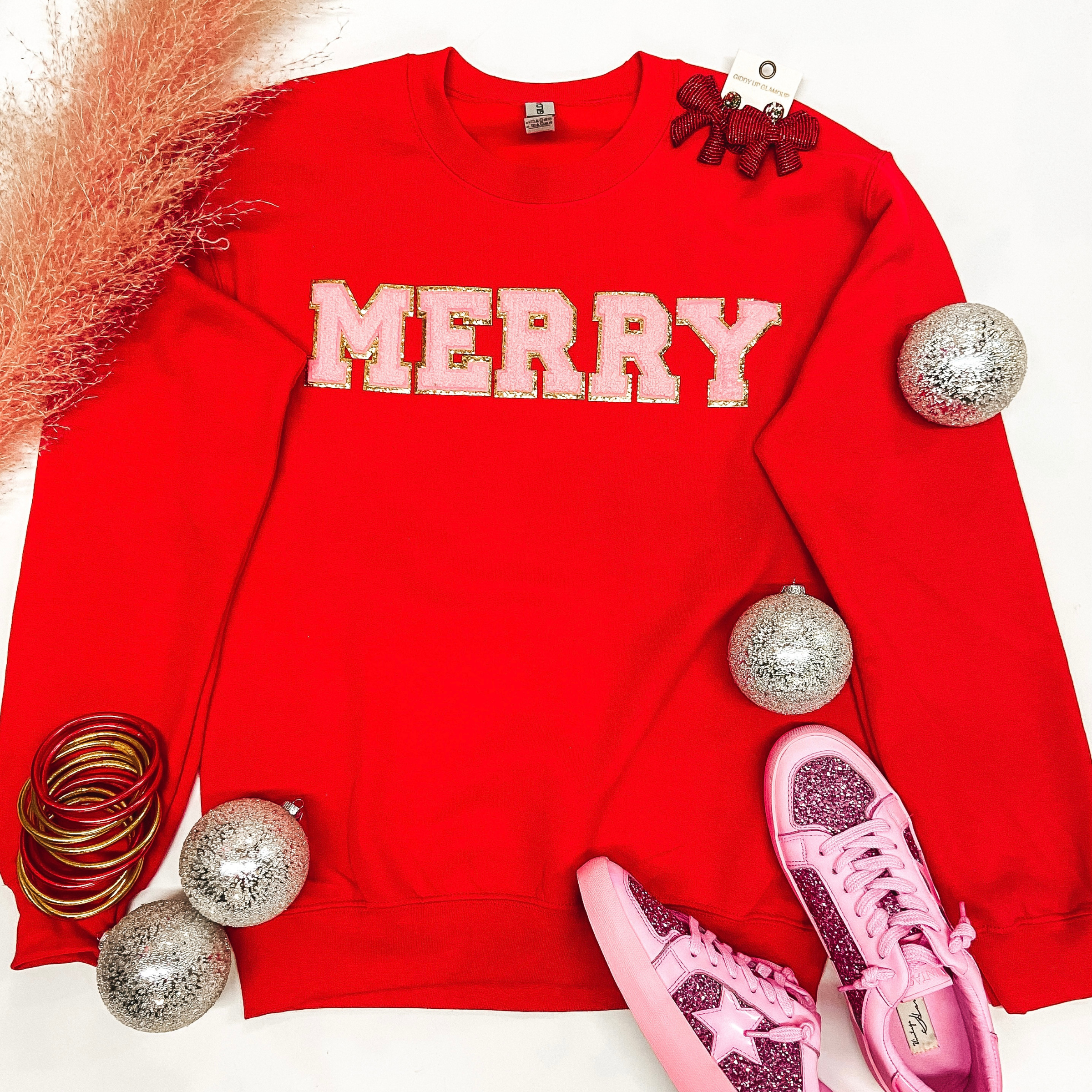 A long sleeve crew neck sweatshirt that is bright green with "Merry" across the chest in pink chenille letters with gold trim. Pictured on a white background with sneakers, gold and pink jewelry, and Christmas ornaments.