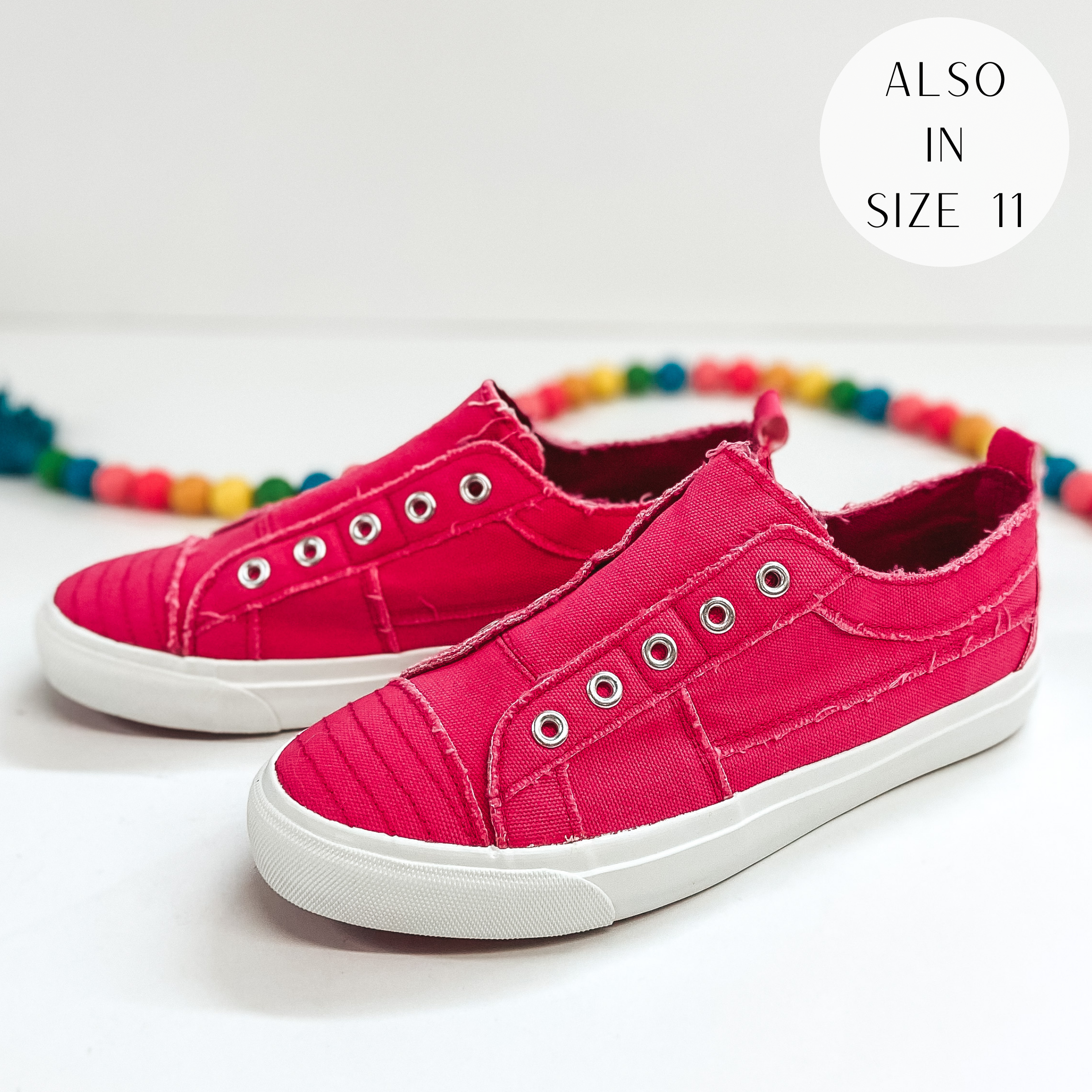 A pair of hot pink sneakers that do not have laces. Pictured on white background with colorful beads.