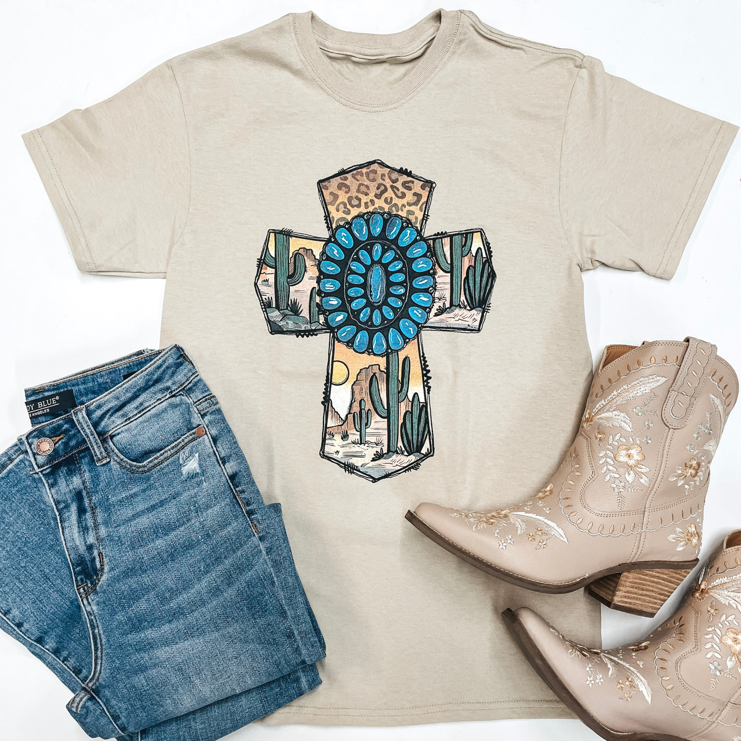 A beige tee is in the center of the picture on top of a white background. In the center of the tee is a cross, outlined in barbwire, with a desert scene inside of it. A turquoise stone sits in the center of the cross. Light wash jeans are on the bottom left side of the picture and cream color boots are on the right. 