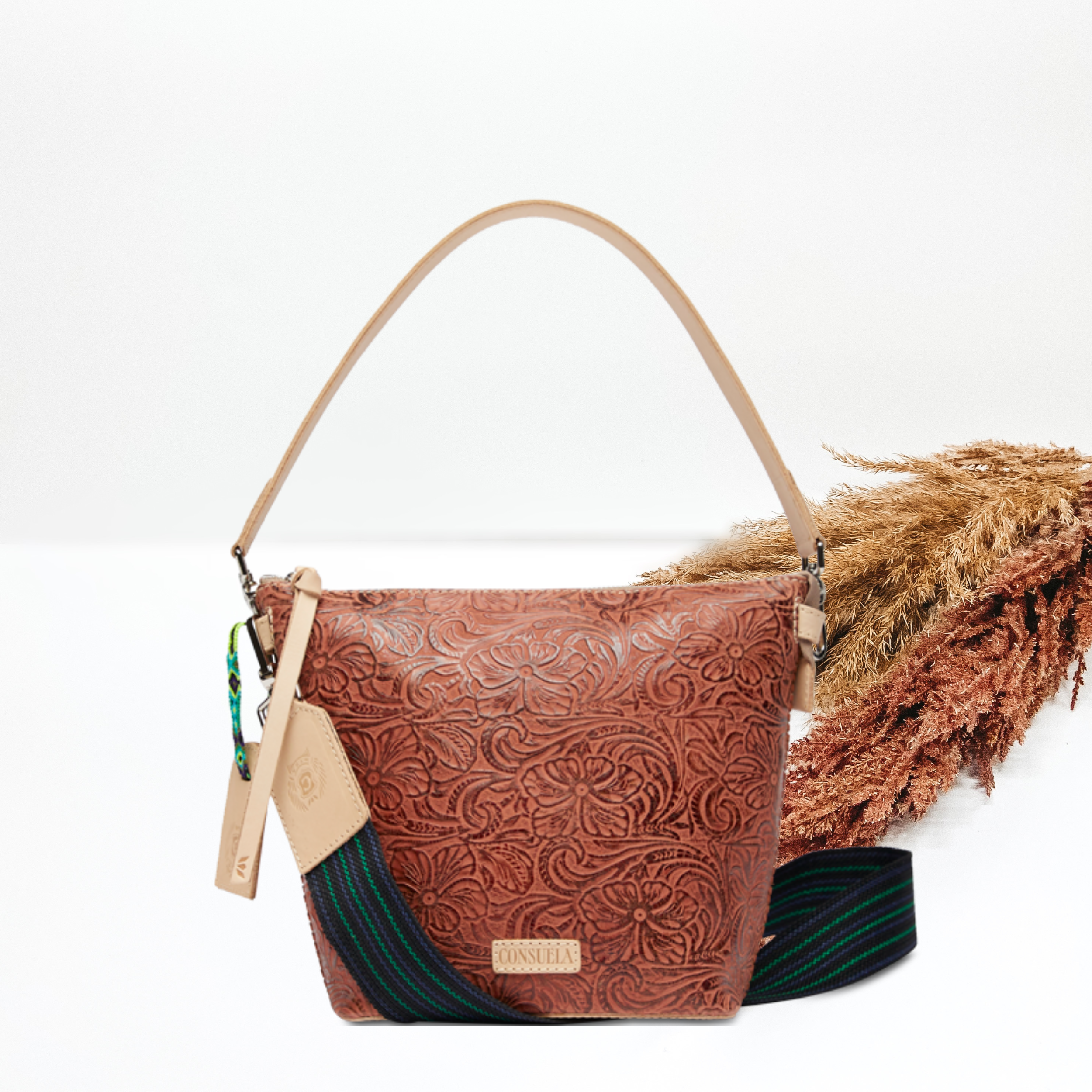 A wedge tooled bag sits in the middle of the picture in a brown tooled pattern. Background is solid white with pompas grass to the right of the purse.  