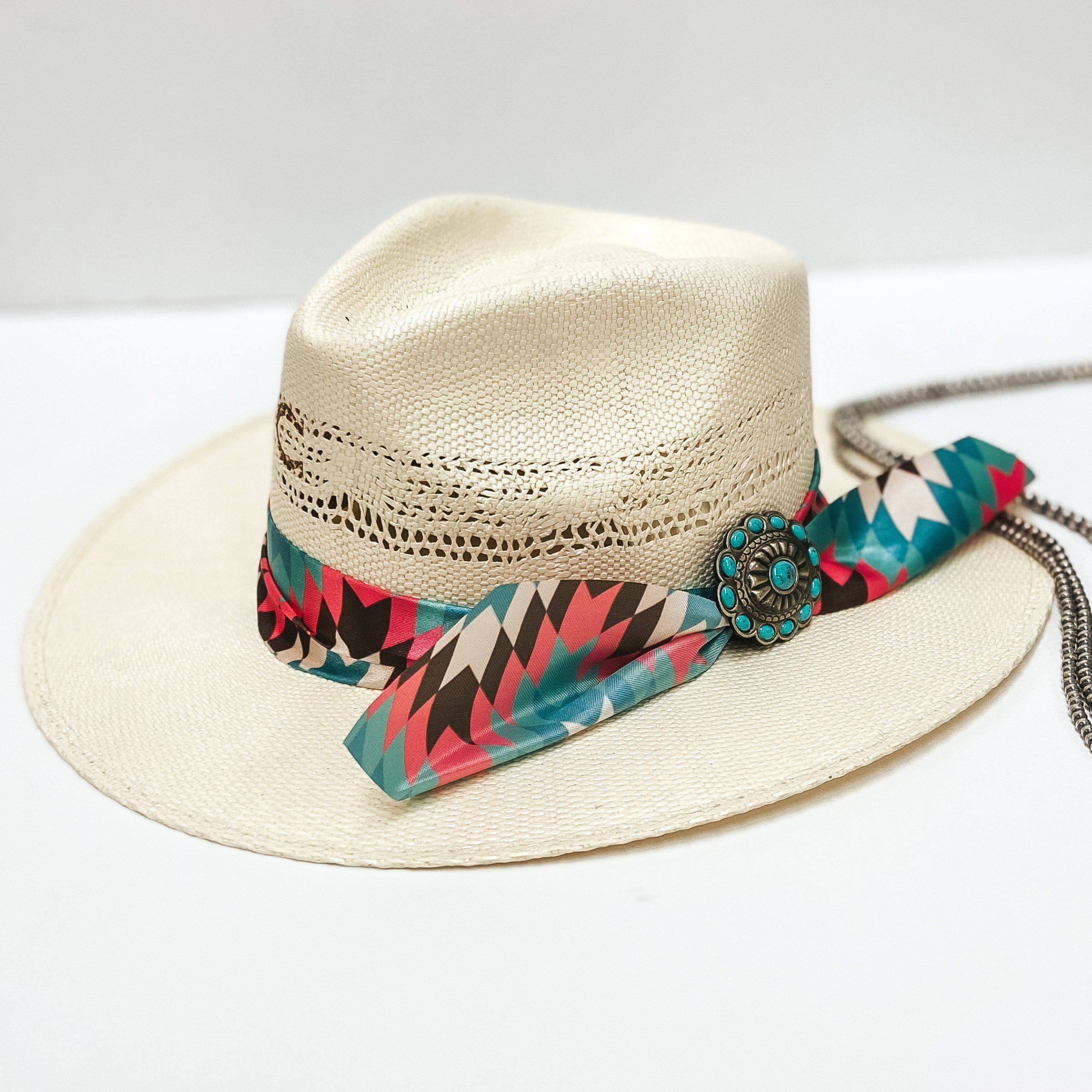 A light beige straw hat with a turquoise and pink ribbon band and a turquoise and silver concho. Pictured on white background with navajo pearls.