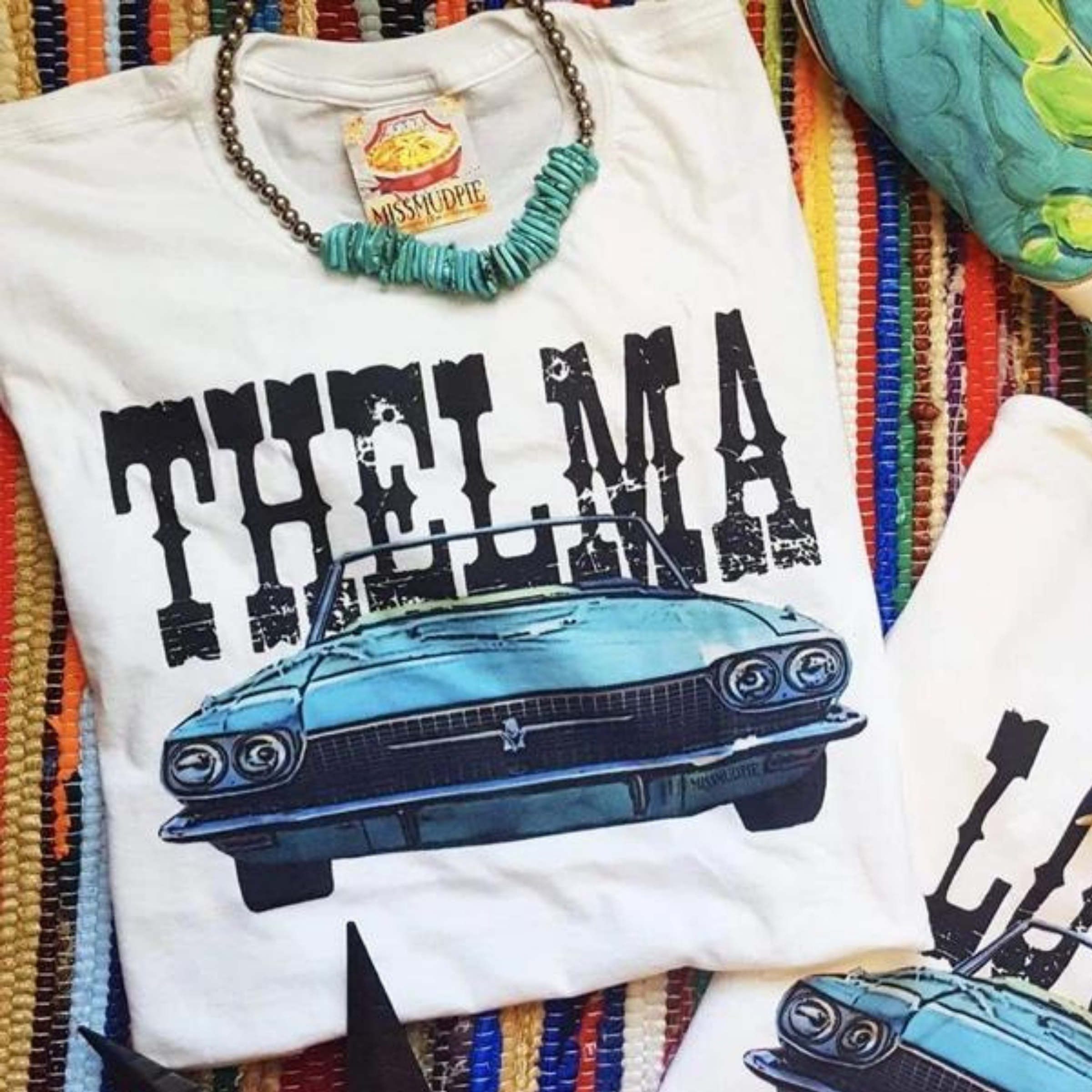 A white tee shirt folded into a rectangle exposing the vintage car graphic that says "Thelma" above. Pictured with a turquoise necklace on a serape rug.