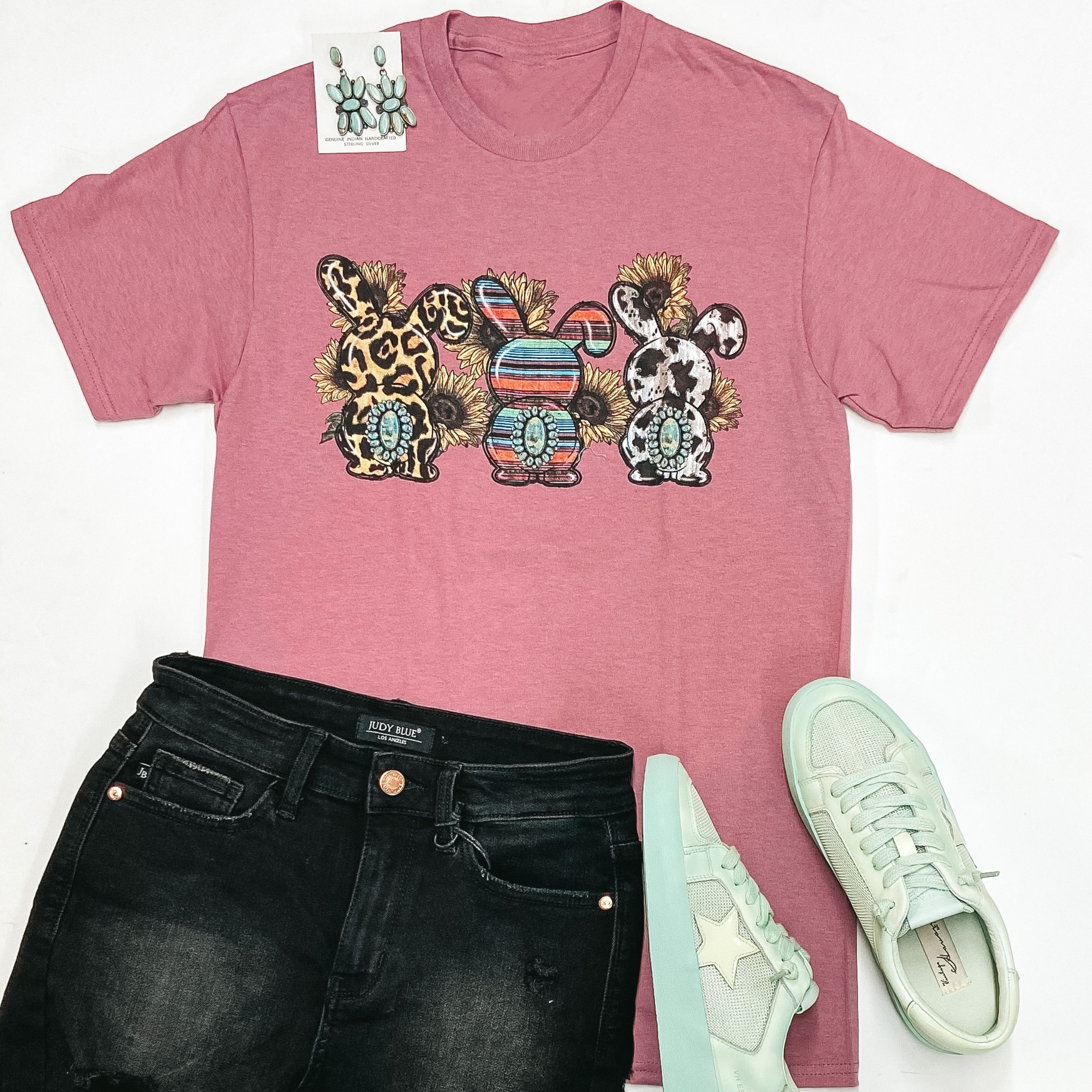 A mauve pink short sleeve graphic tee with a leopard print, serape print, and cow print bunny with sunflowers around them. This tee is pictured on a white background with black shorts, mint green sneakers, and genuine turquoise jewelry.