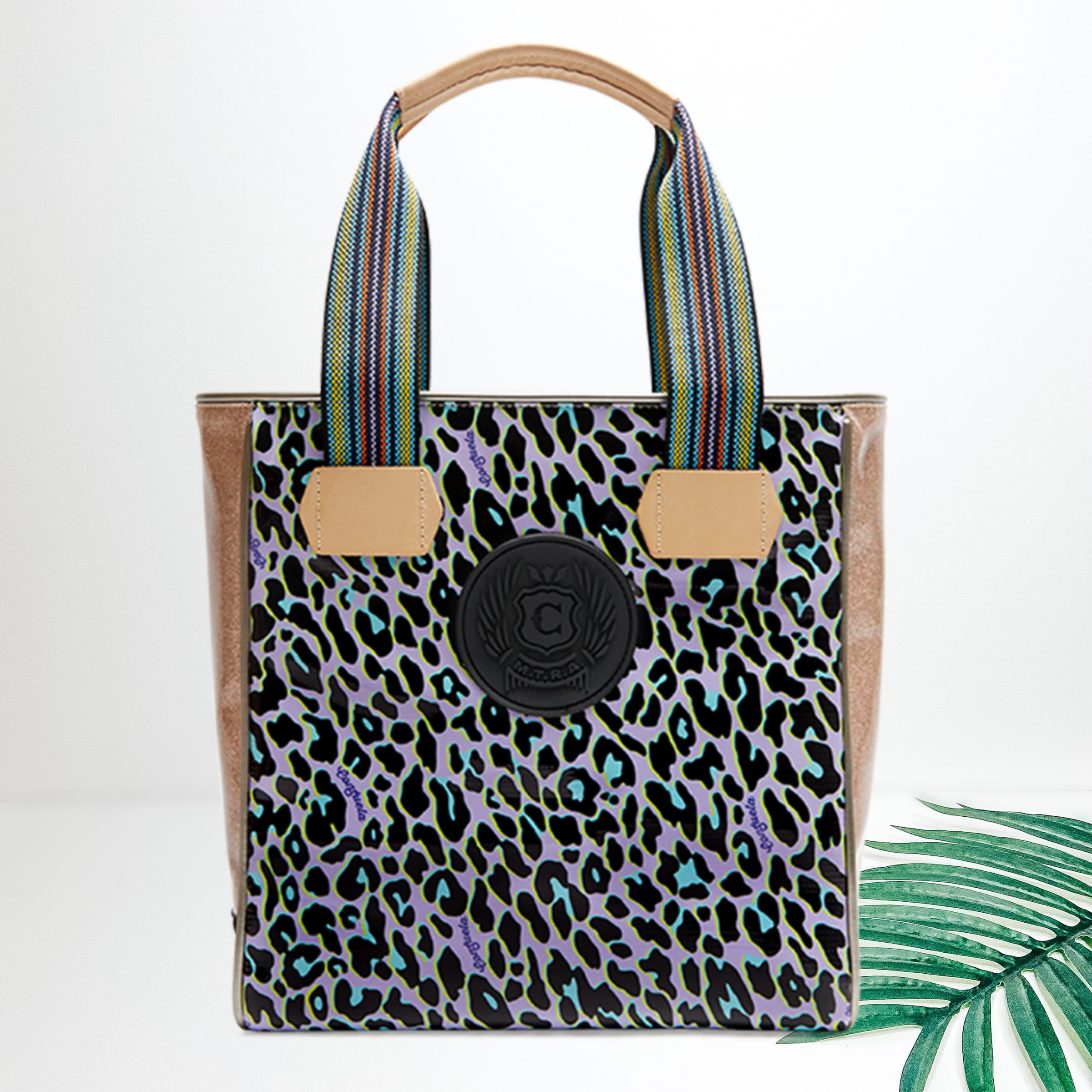A green and purple leopard print bag with a black patch. Pictured on white background with a palm leaf.
