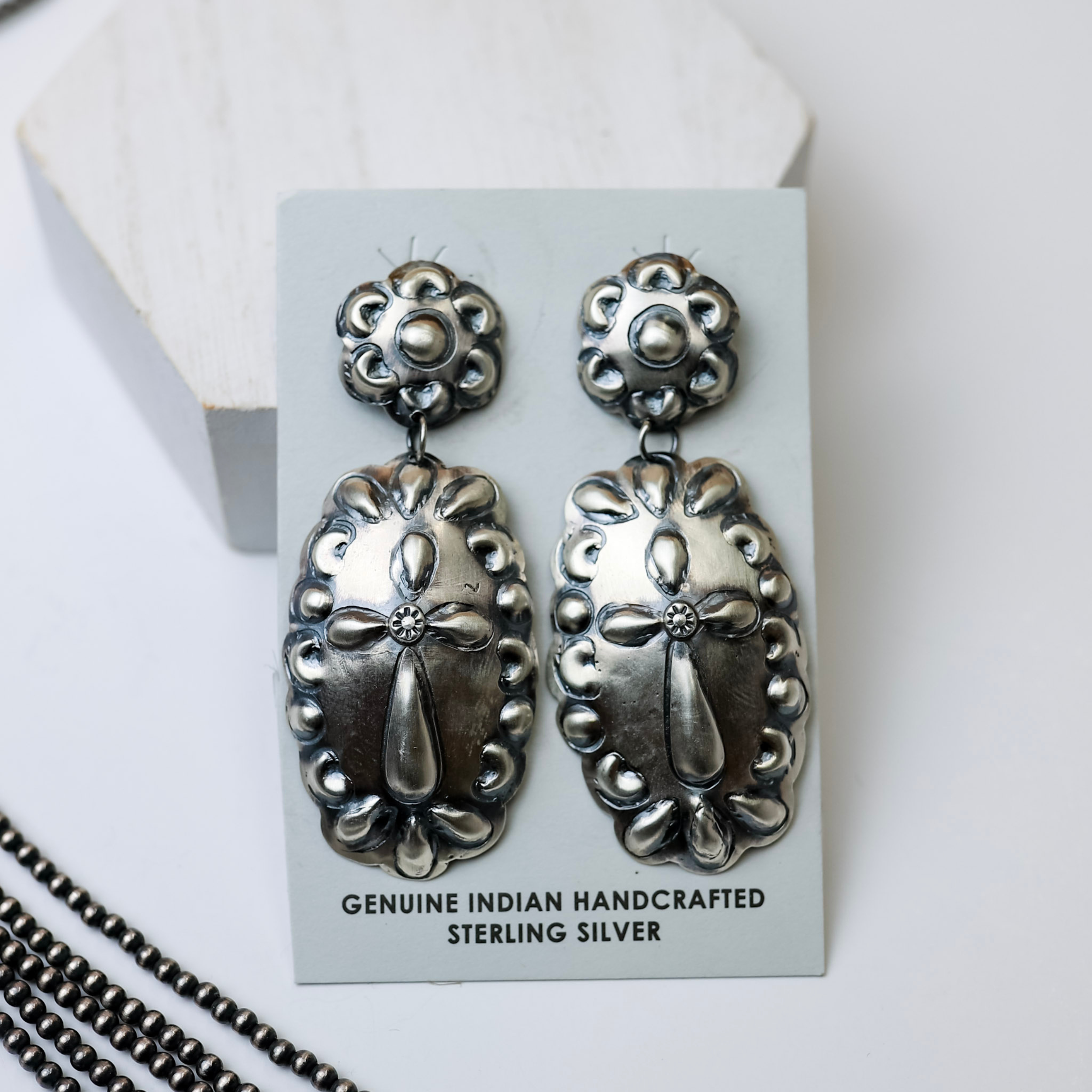 Tim Yazzie | Navajo Handmade Sterling Silver Flower Concho Post Stud and Dangle Earrings with Cross are laid in the center of the picture. Navajo pearls are to the left of the earrings, all on a white background. 