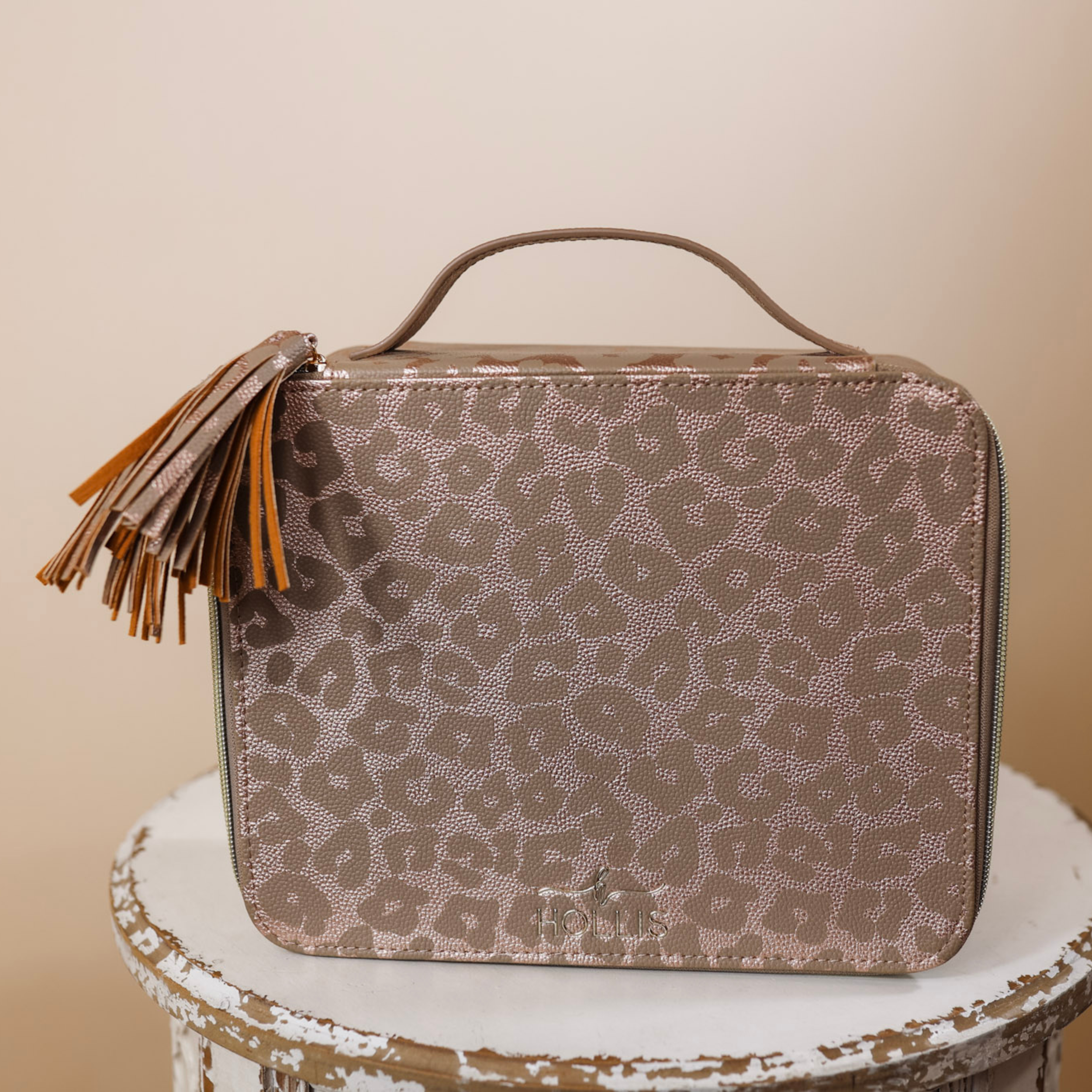 A metallic leopard bag with tassels and a handle is sitting in the middle of the picture. Background is solid tan. 