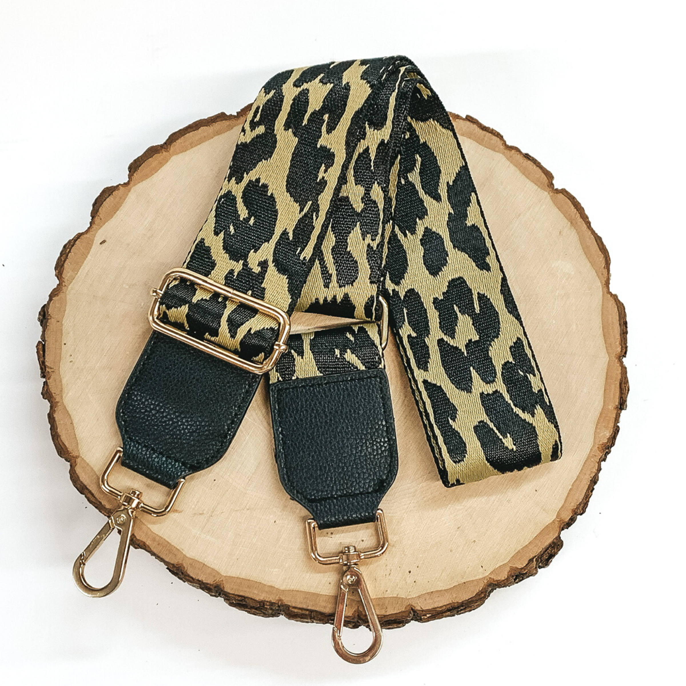 Gold purse strap with a black leopard print. This purse strap includes gold accents and black pieces at each end. This purse strap is pictured on a piece of wood on a white background. 