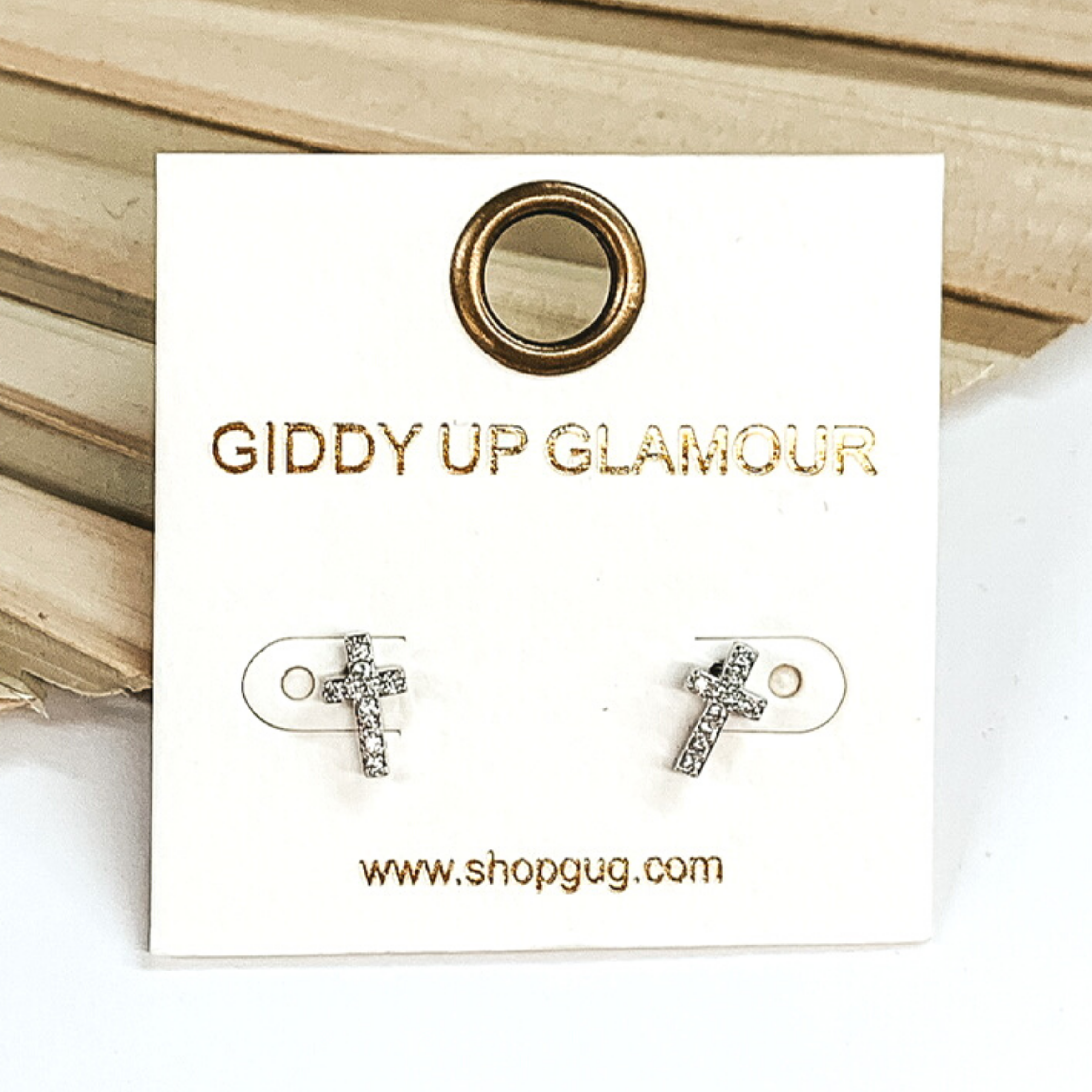 Small, silver cross stud earrings with clear crystals. These earrings are pictured on a white earrings card on a white and brown background.