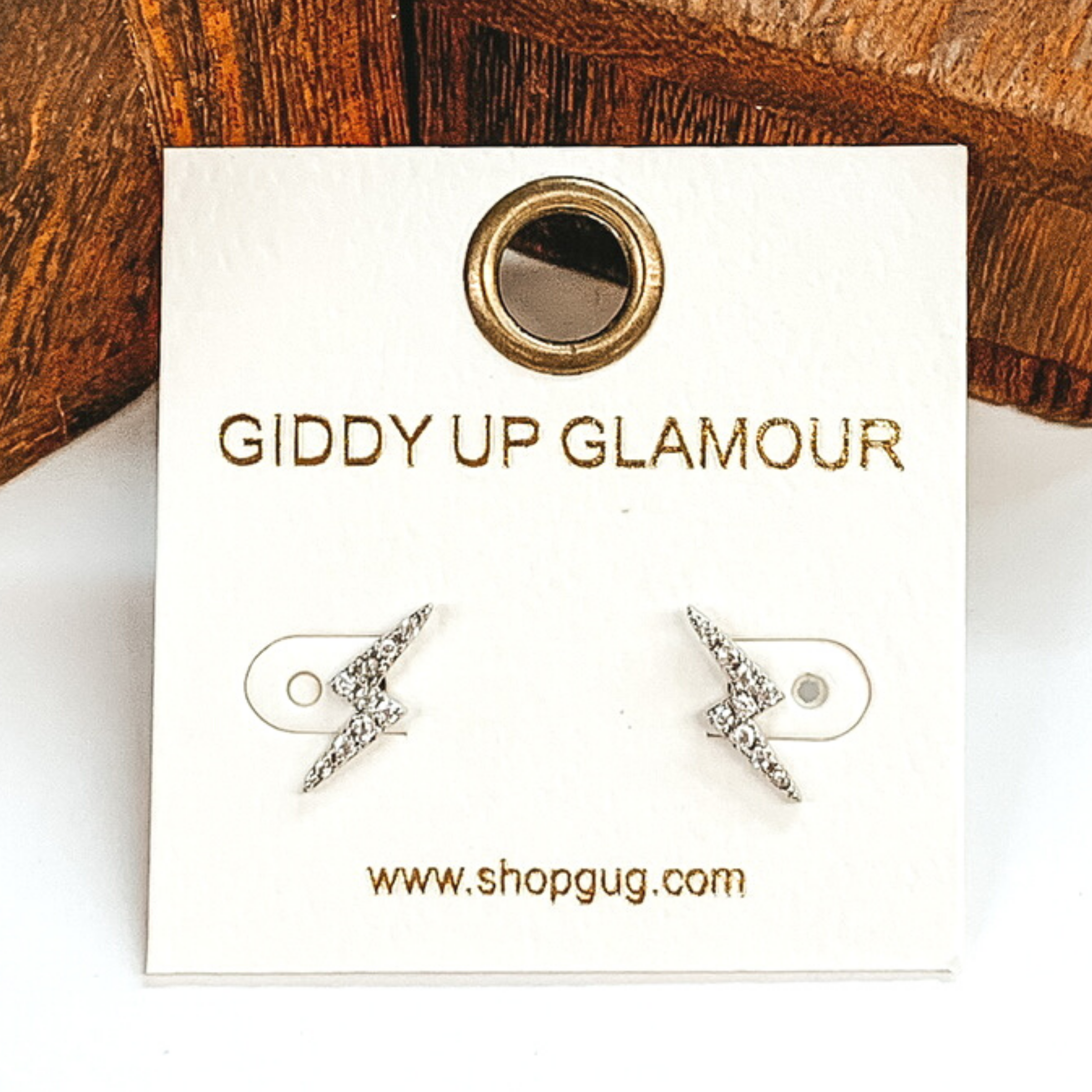 Small, silver thunder bolt stud earrings with clear crystals. These earrings are pictured on a white earrings card on a white and brown background.