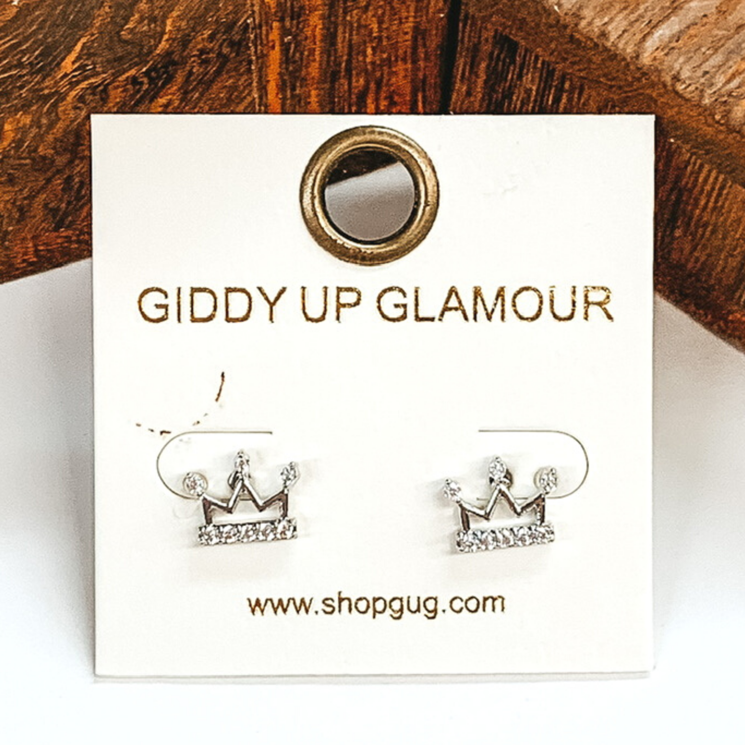 Small, silver crown outline stud earrings with clear crystals. These earrings are pictured on a white earrings card on a white and brown background.