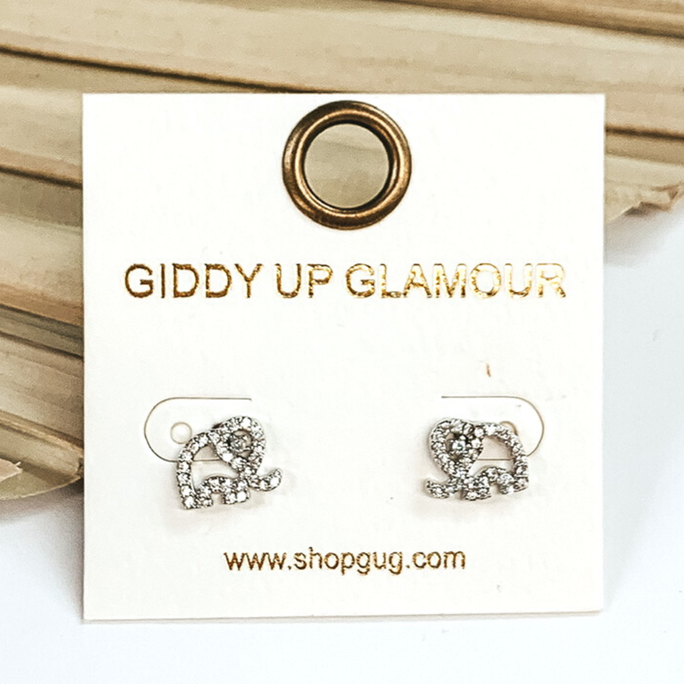 Small, silver elephant outline stud earrings with clear crystals. These earrings are pictured on a white earrings card on a white and green background. 
