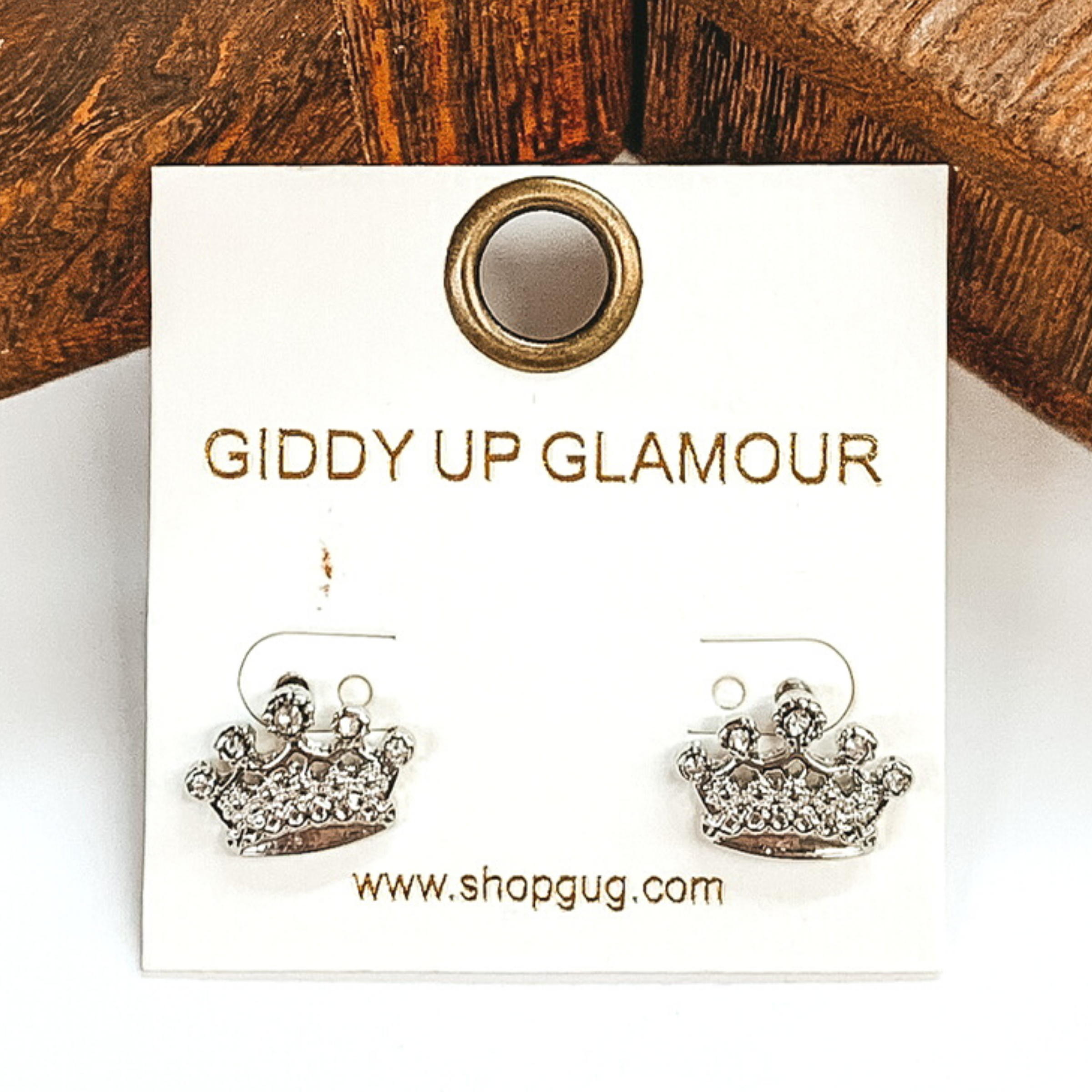 Silver crown stud earrings with clear crystals. These earrings are pictured on a white and brown background. 
