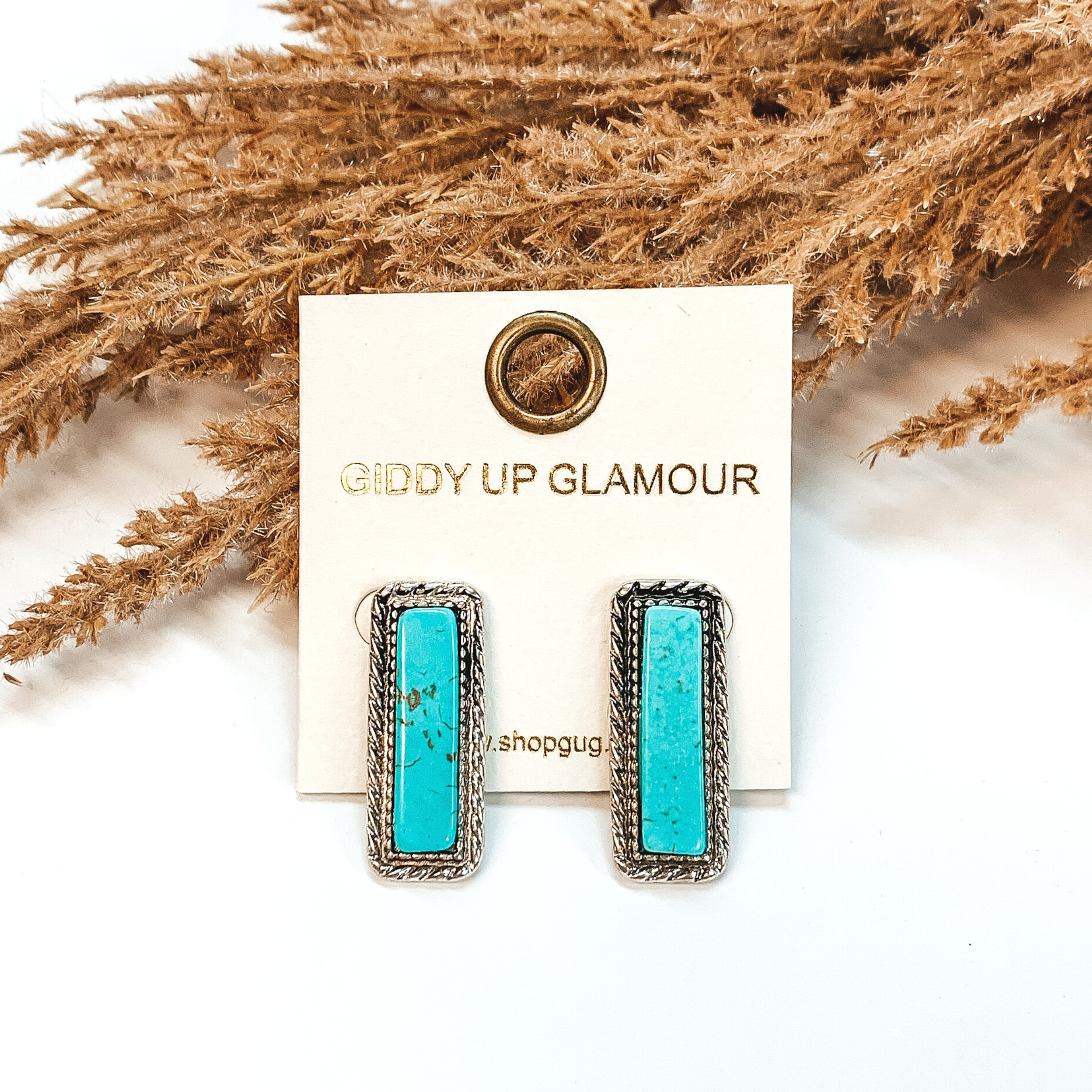 Silver rectangle stud earrings with a turquoise rectangle stone inlay. These earrings are pictured on a white background in front of brown floral decor. 