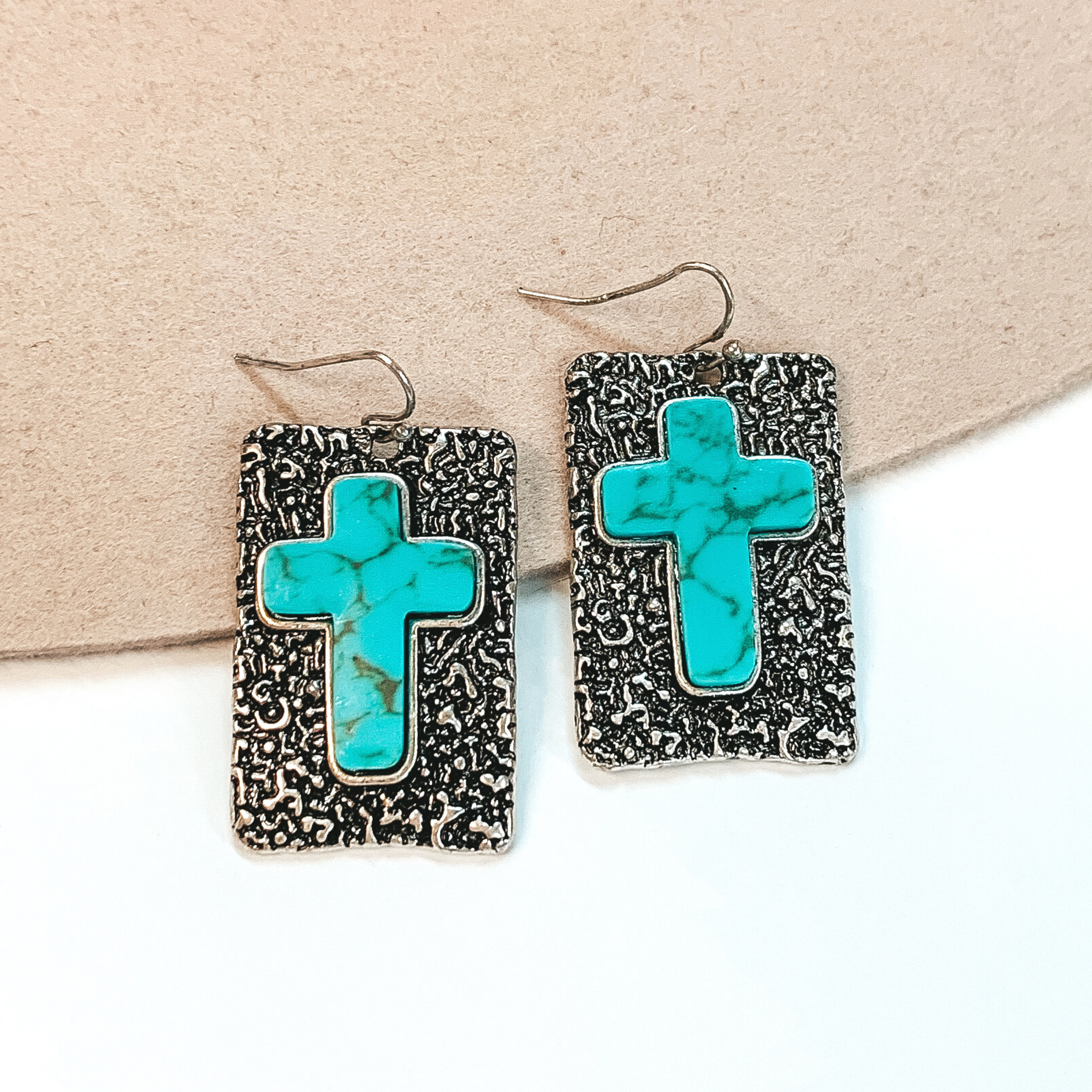Fish hook earrings with silver rectangle pendant that has engraving detailing. In the center of the pendant, there is a turquoise cross stone. These earrings are pictured on a white and beige background. 
