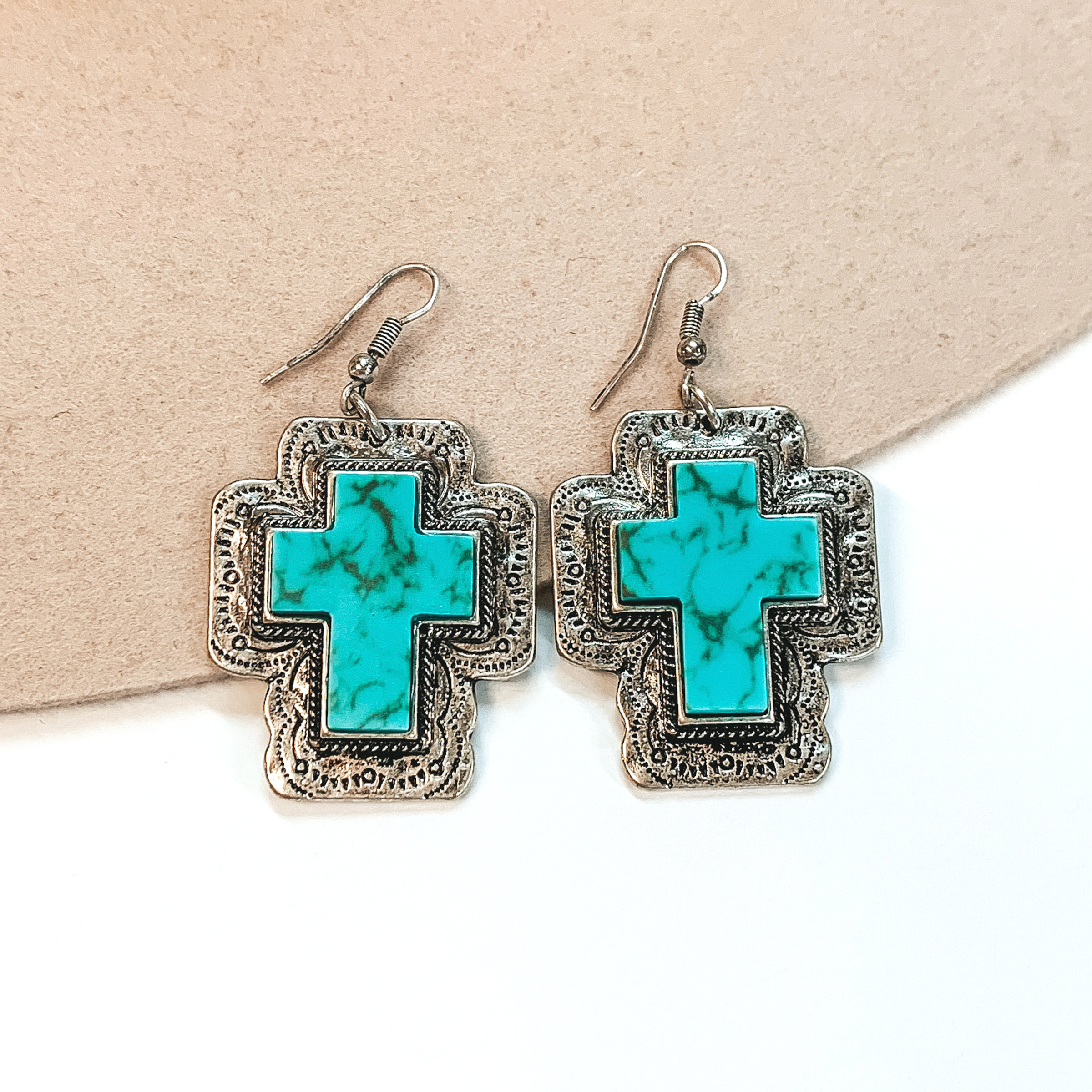 Fish hook earrings with silver cross pendant with engraving detailing. In the center of the pendant, there is a turquoise cross stone. These earrings are pictured on a white and beige background. 