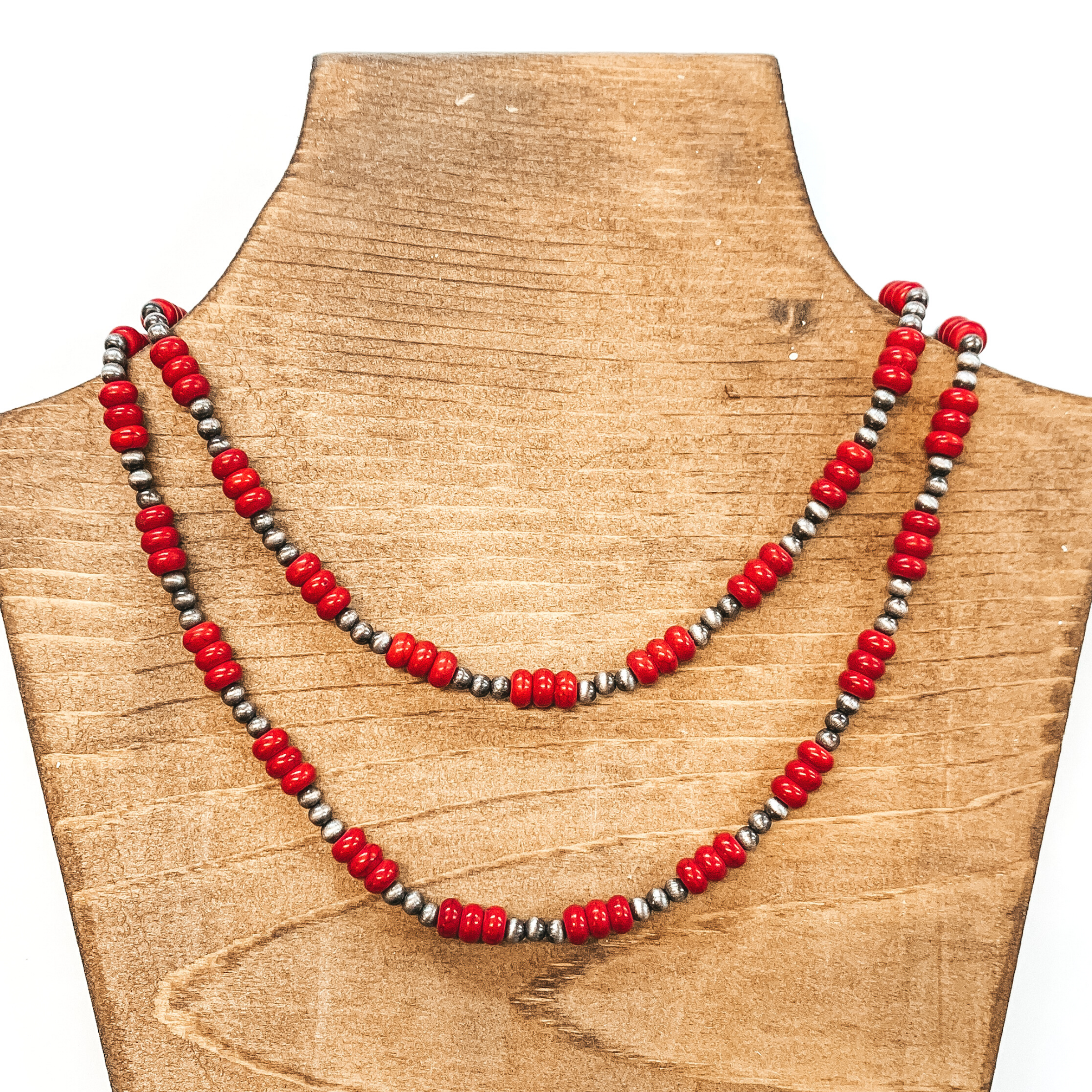 Two strand necklace that includes silver bead segments with bigger, red bead segments. This necklace is pictured on a brown necklace holder on a white background.