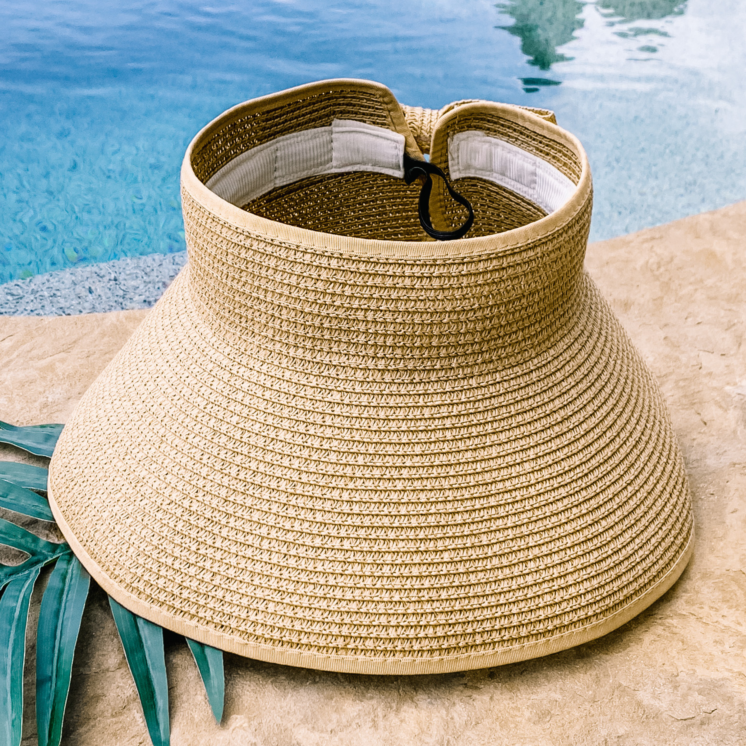 Natural, woven velcro visor that is pictured with a green leaf under it. This visor is pictured on a a tan rock in front of a pool.