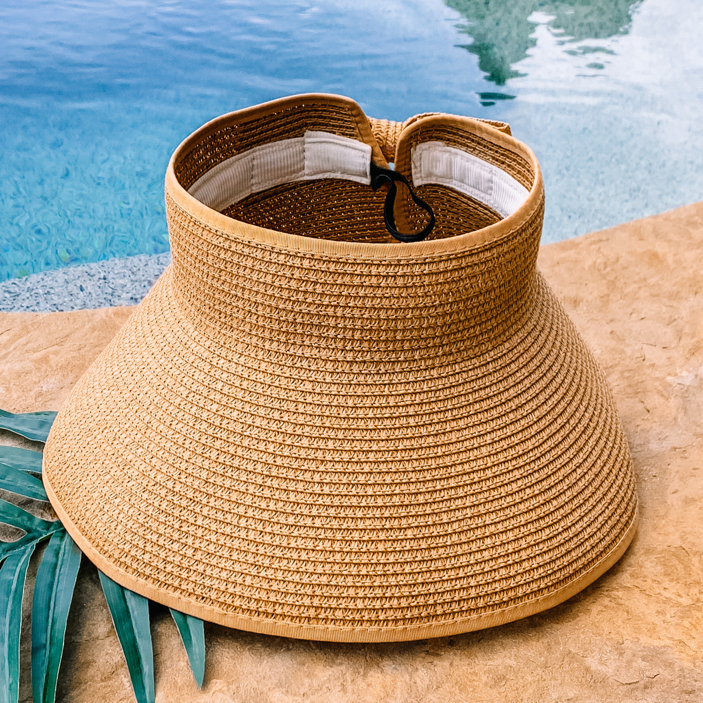 Tan, woven velcro visor that is pictured with a green leaf under it. This visor is pictured on a a tan rock in front of a pool.