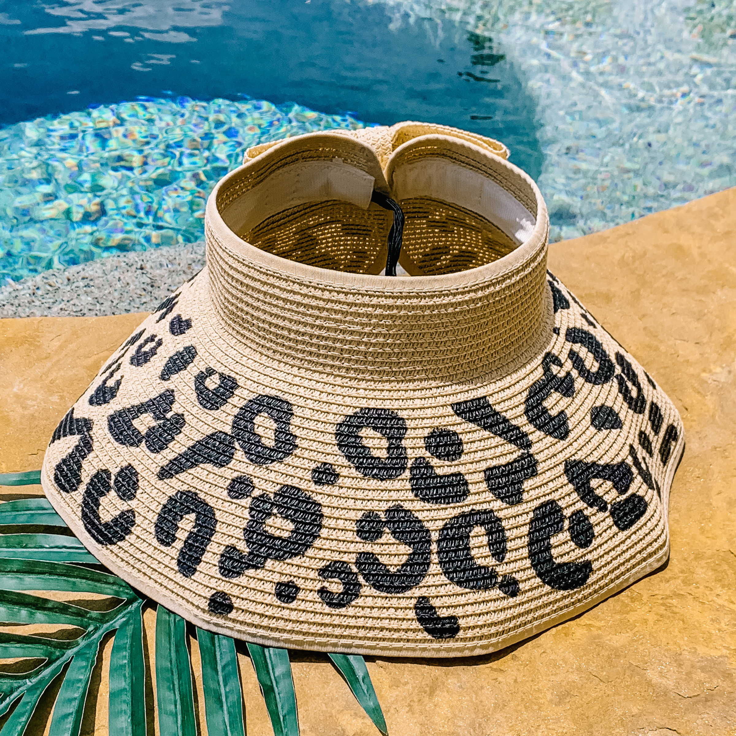 Tan, woven velcro visor with a leopard print that is pictured with a green leaf under it. This visor is pictured on a a tan rock in front of a pool.