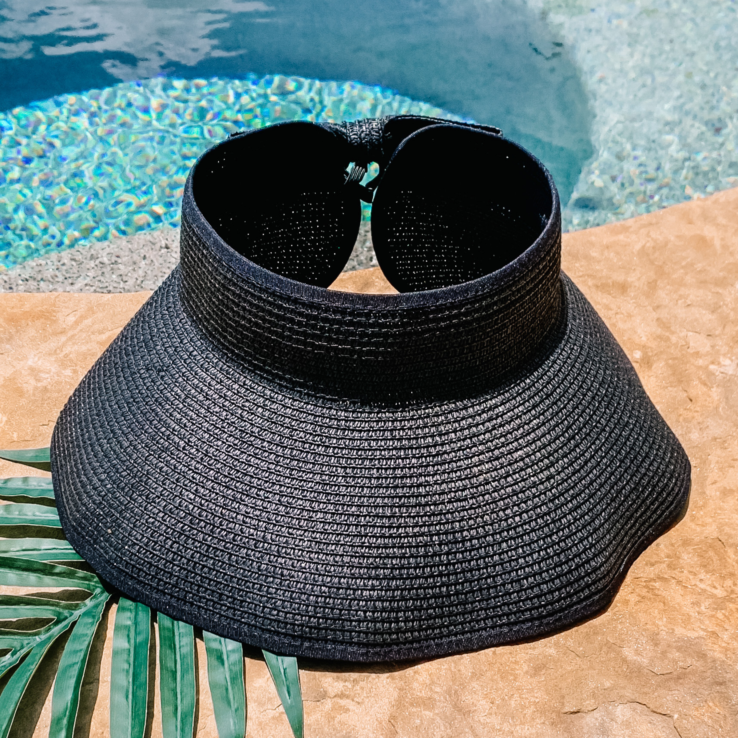 Black, woven velcro visor that is pictured with a green leaf under it. This visor is pictured on a a tan rock in front of a pool.