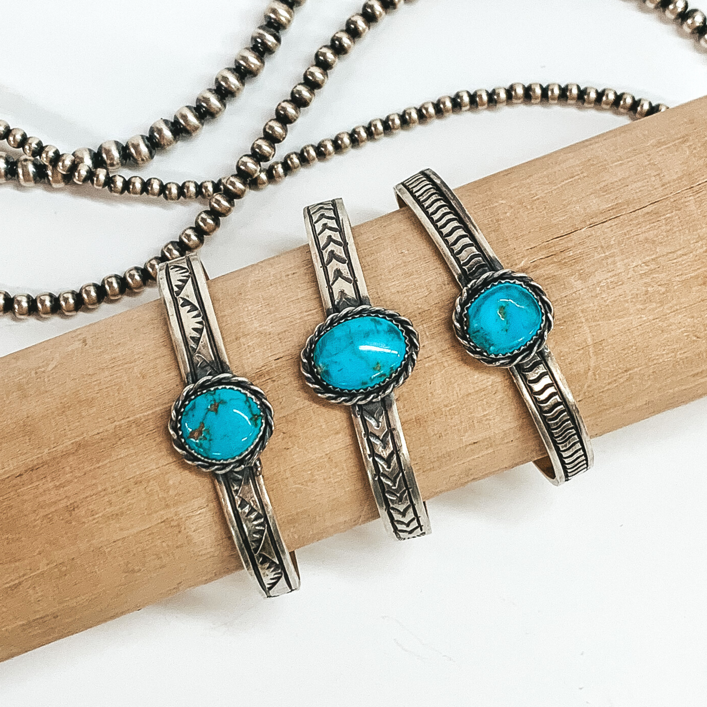 Three silver, engraved bracelets with a center turquoise stone. These bracelet are pictured on a tan bracelet holder on a white background. 