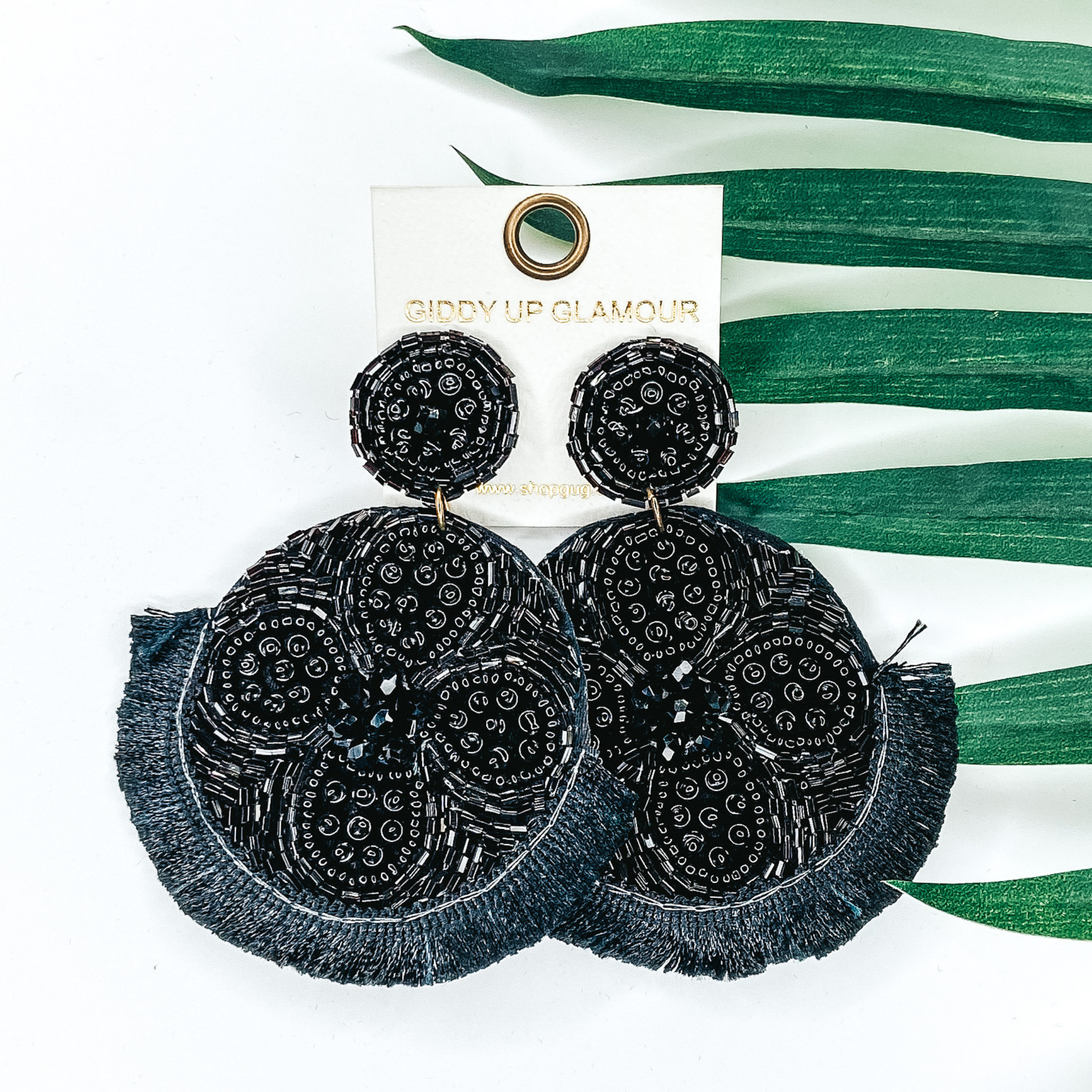 Black beaded circle post back earrings. Hanging from the earrings are black beaded circle pendant with black fringe. These earrings are pictured on a white background with green leaves behind them.