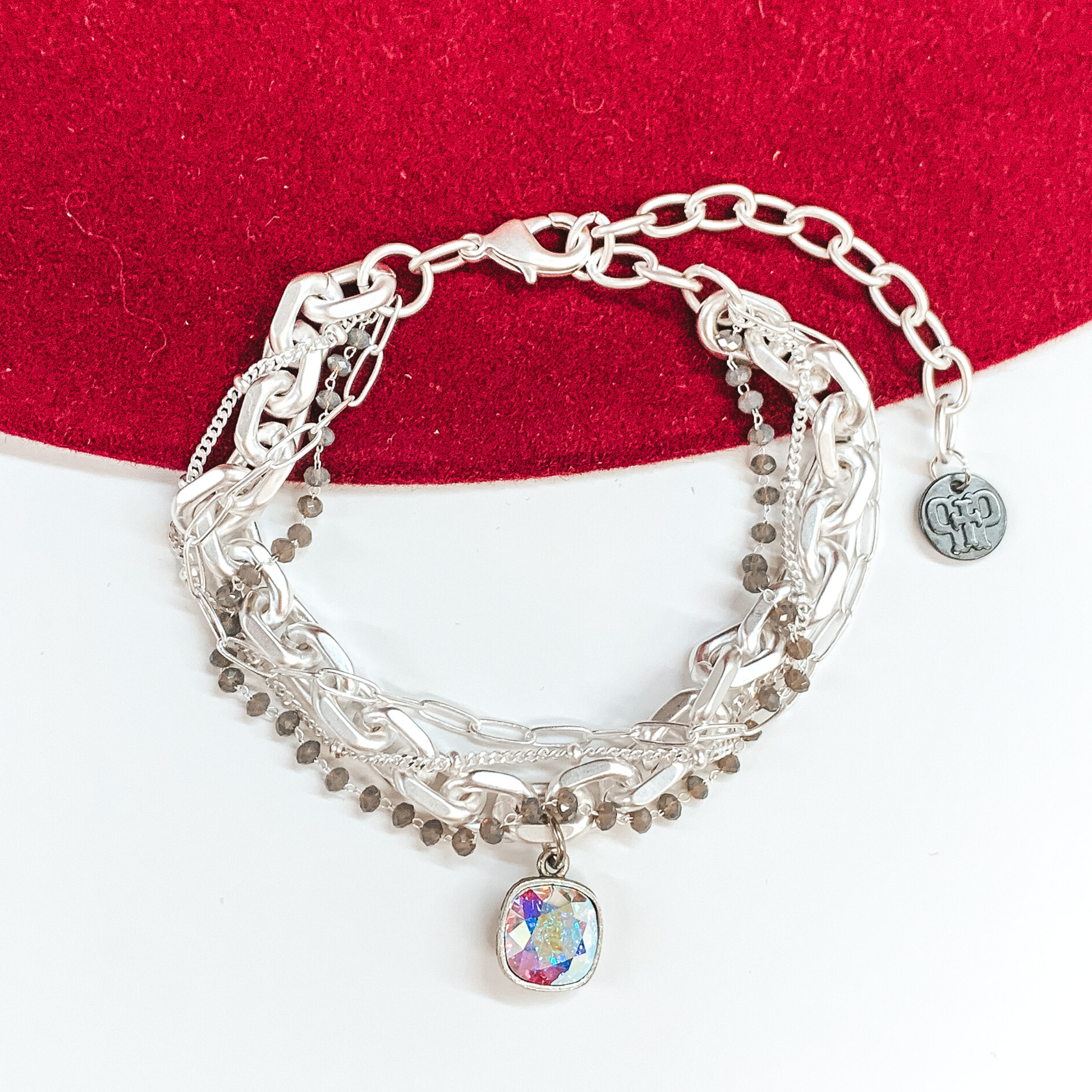 Silver, adjustable bracelet that includes a thick chain strnad, crystal beaded strand, a thin paperlcip chain strand, and a small chain strand. There is also an AB cushion cut crystal drop. this bracelet is pictured on a white and red background.