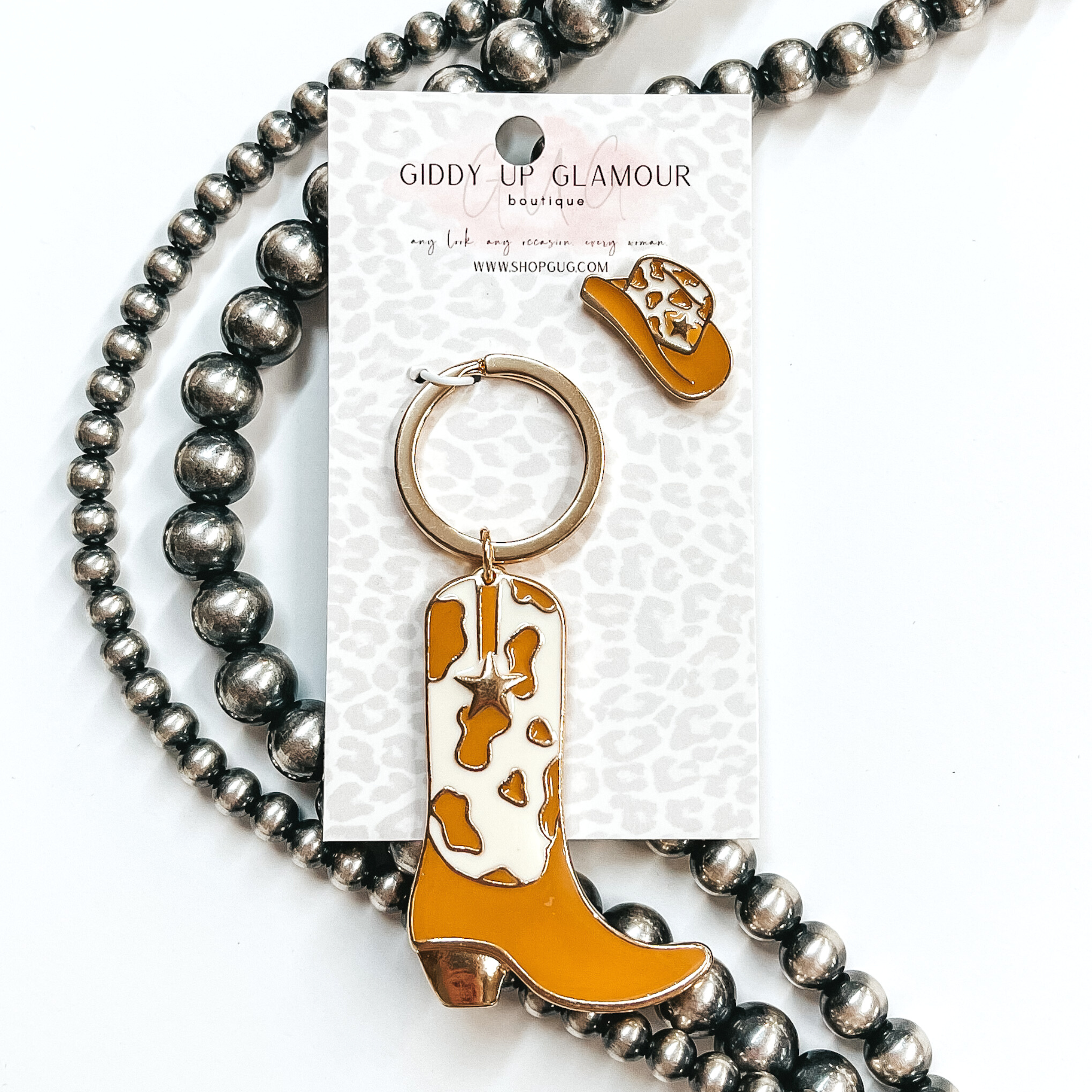 Gold key ring with boot pendant that has a tan and ivory cow print design. The boot also has a gold star. There is also a white and pink hat pin. These two are pictured on a white cardstock on a white background with silver beads. 