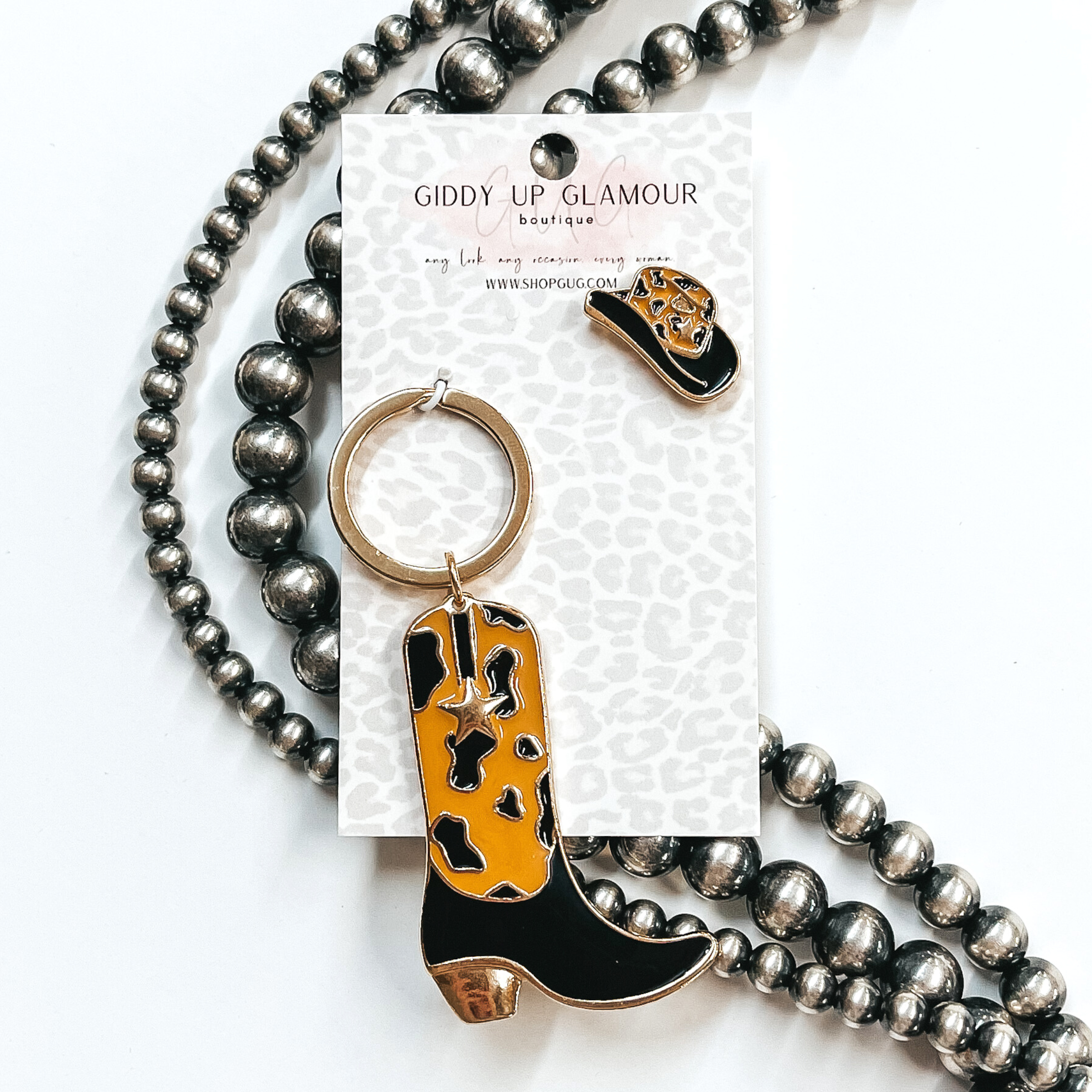 Gold key ring with boot pendant that has a black and tan cow print design. The boot also has a gold star. There is also a white and pink hat pin. These two are pictured on a white cardstock on a white background with silver beads. 