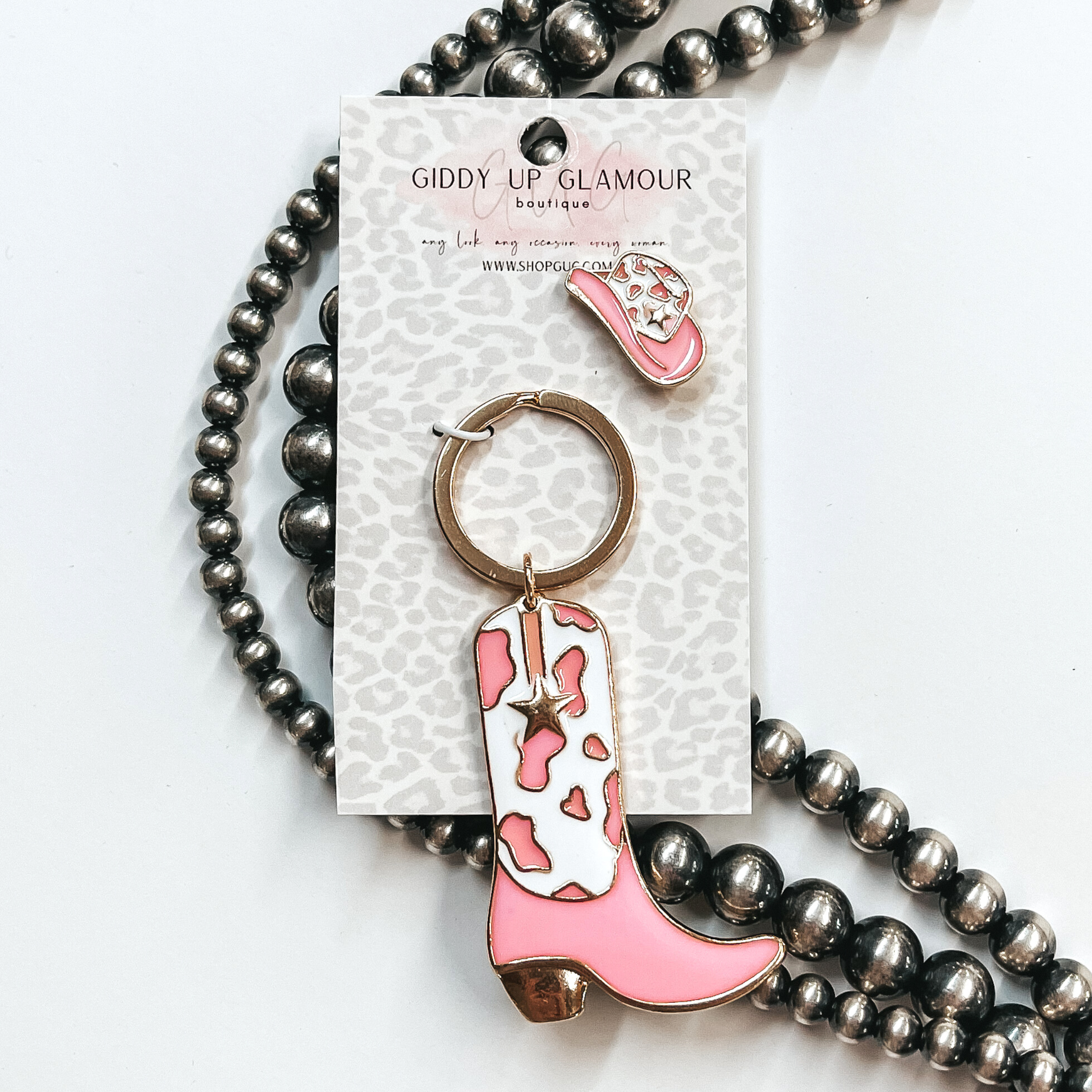 Gold key ring with boot pendant that has a white and pink cow print design. The boot also has a gold star. There is also a white and pink hat pin. These two are pictured on a white cardstock on a white background with silver beads. 