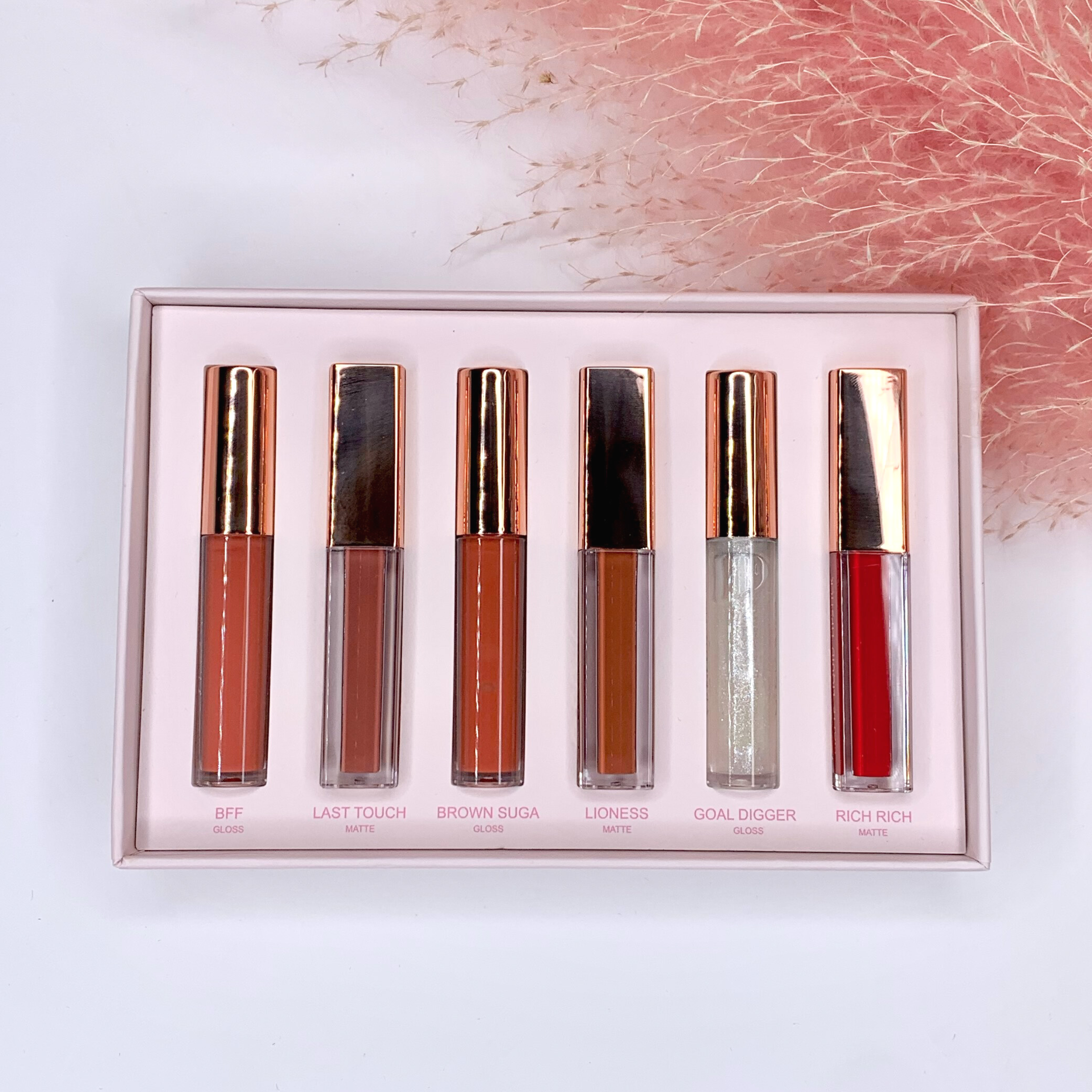 In a light pink box, there are six lip gloss tubes. Each lip gloss has its shade written under the tube. This set is pictured on a white background with pink pompus grass in the top right corner. 