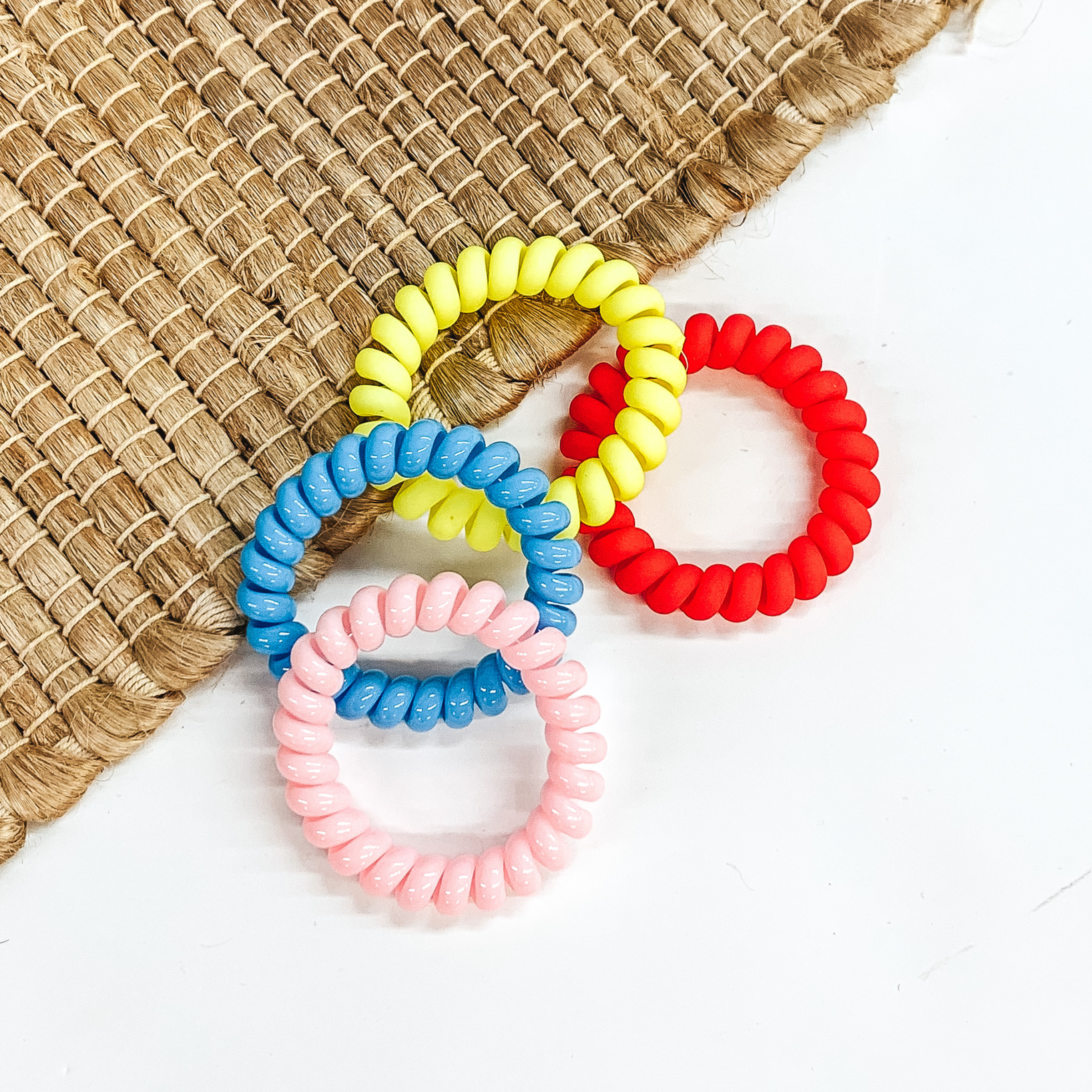 Four spiral scrunchies in the colors light pink, blue, yellow, and coral red pictured on a white and basket weave material background. 
