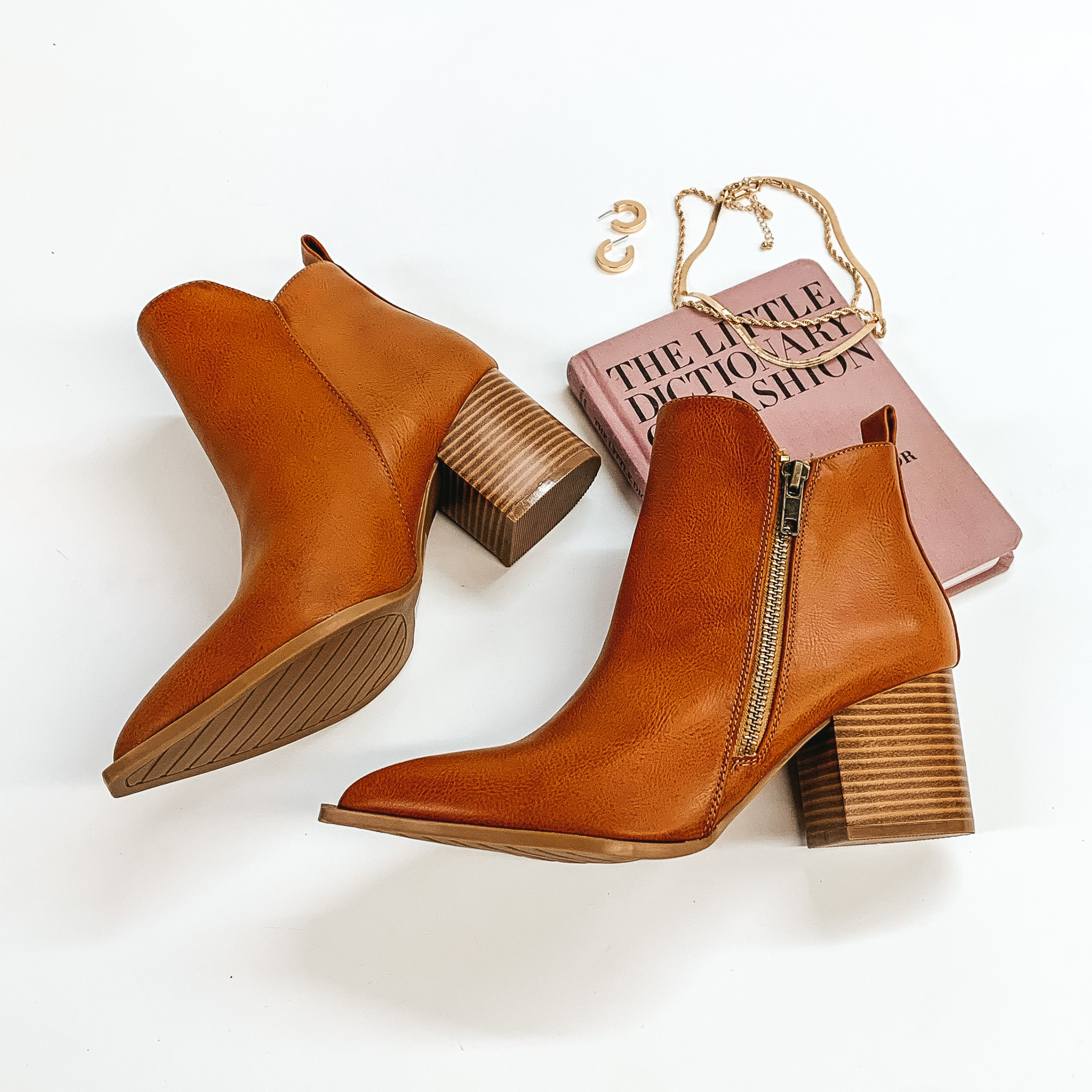 Ankle booties in cognac with a side zipper. These booties also have a wooden heel. These booties are pictured on a white background with gold jewelry and a book.  