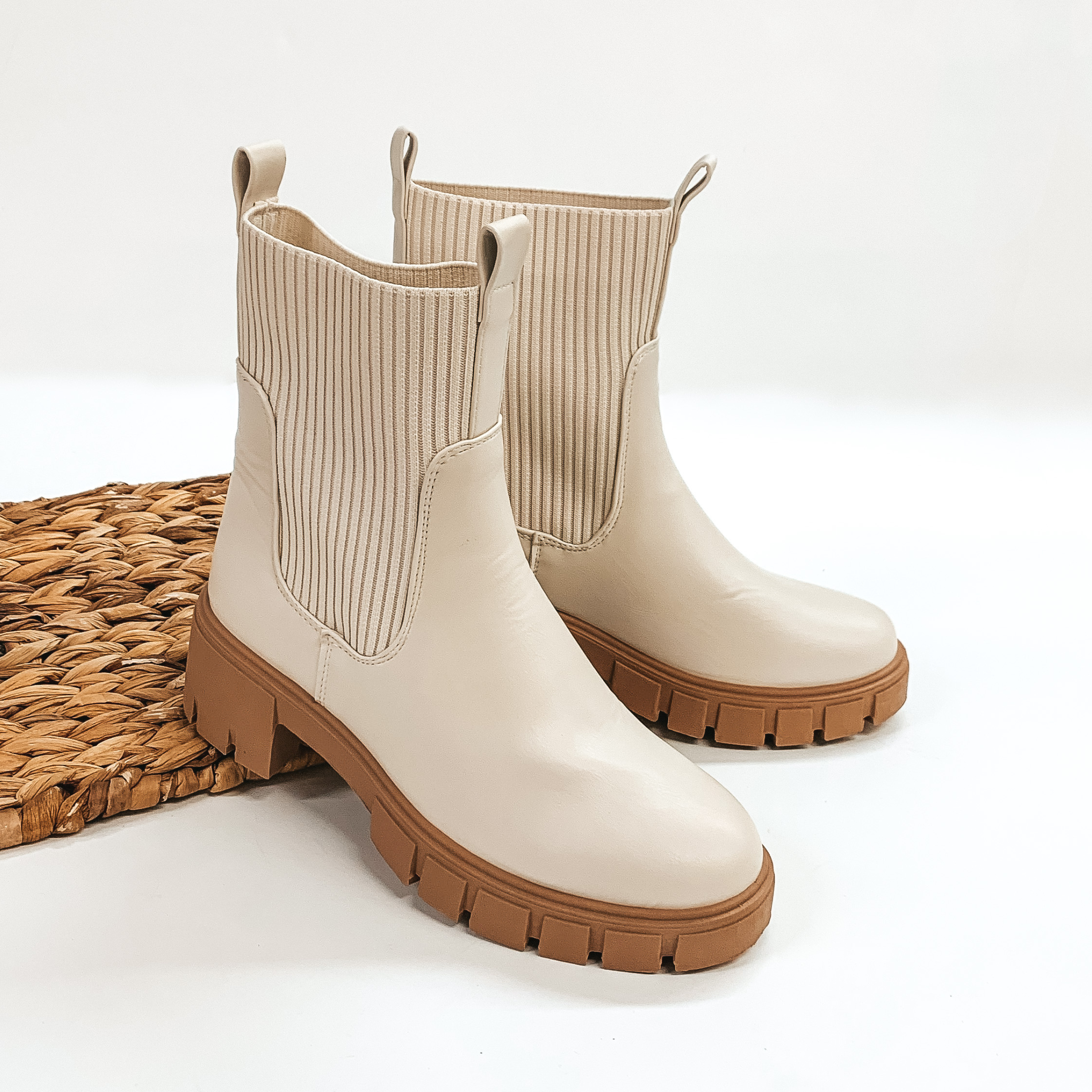 Ankle high off white colored slip on bootie with a tan sole. The sides are elastic and also have a front and back pull on tab. These booties are pictured propped on a basket weave material on a white background. 