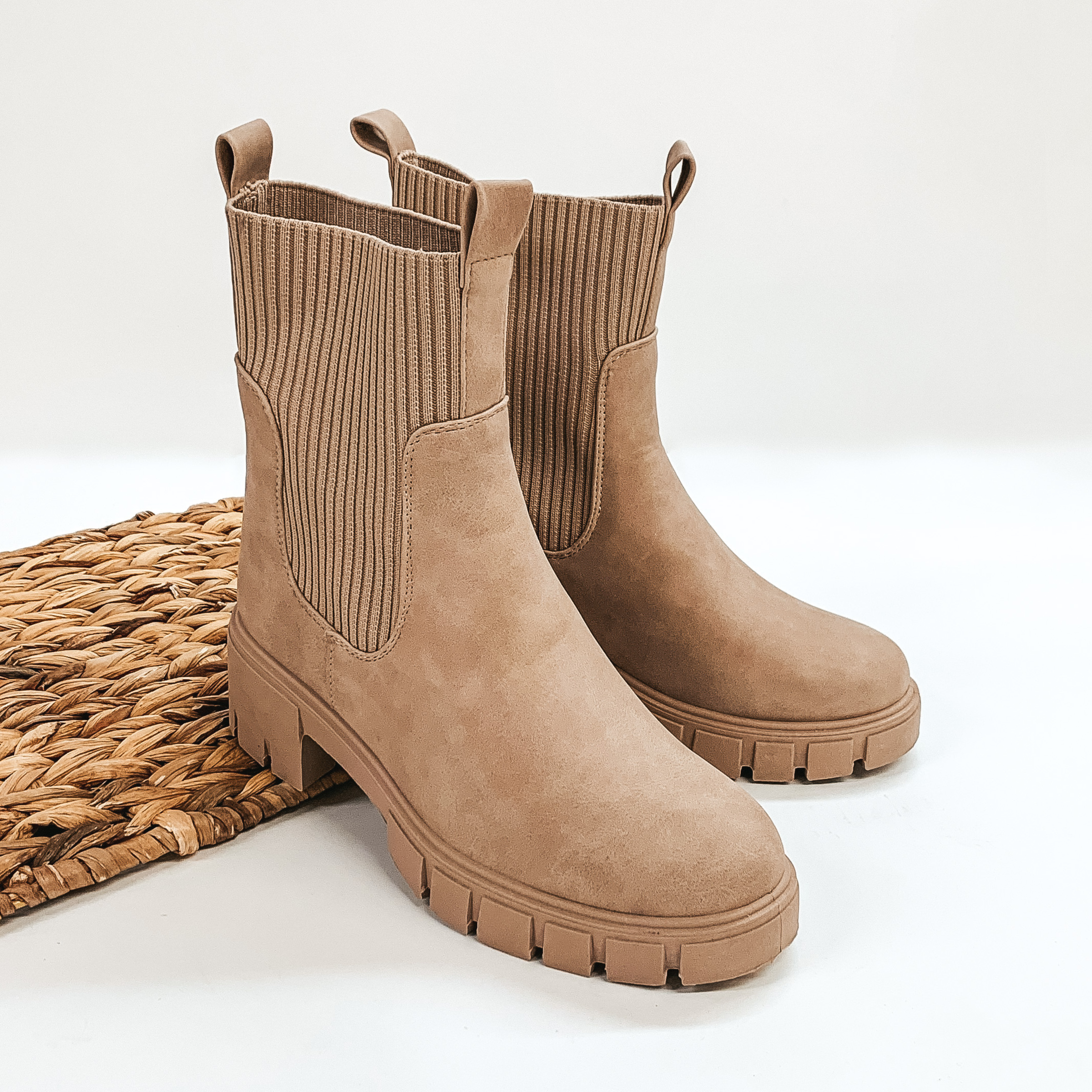 Ankle high taupe colored slip on bootie. The sides are elastic and also have a front and back pull on tab. These booties are pictured propped on a basket weave material on a white background. 