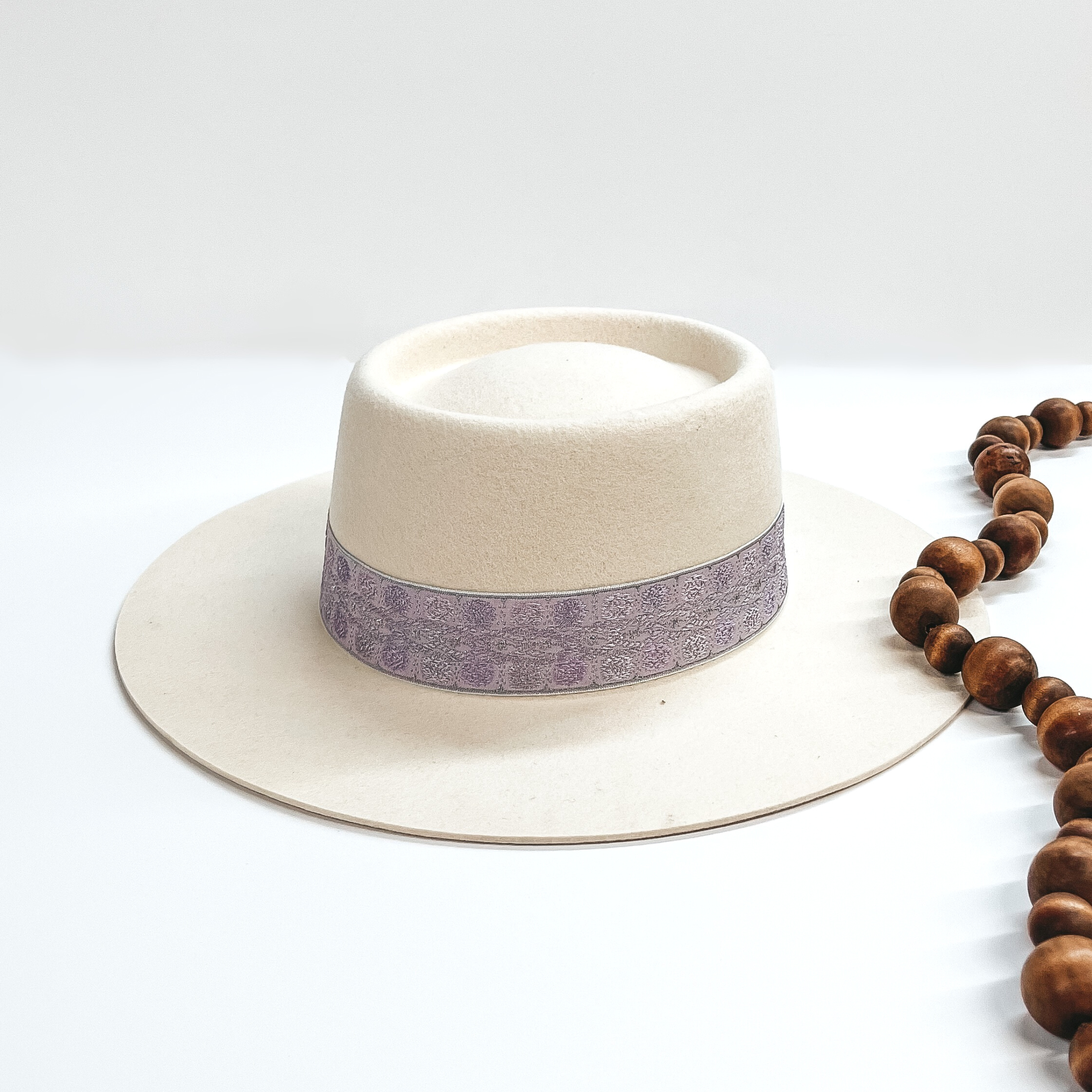 Ivory felt hat with an oval crown and flat brim. This hat also has a light purple lace hat band. This hat is pictured on a white background with brown decorative beads. 