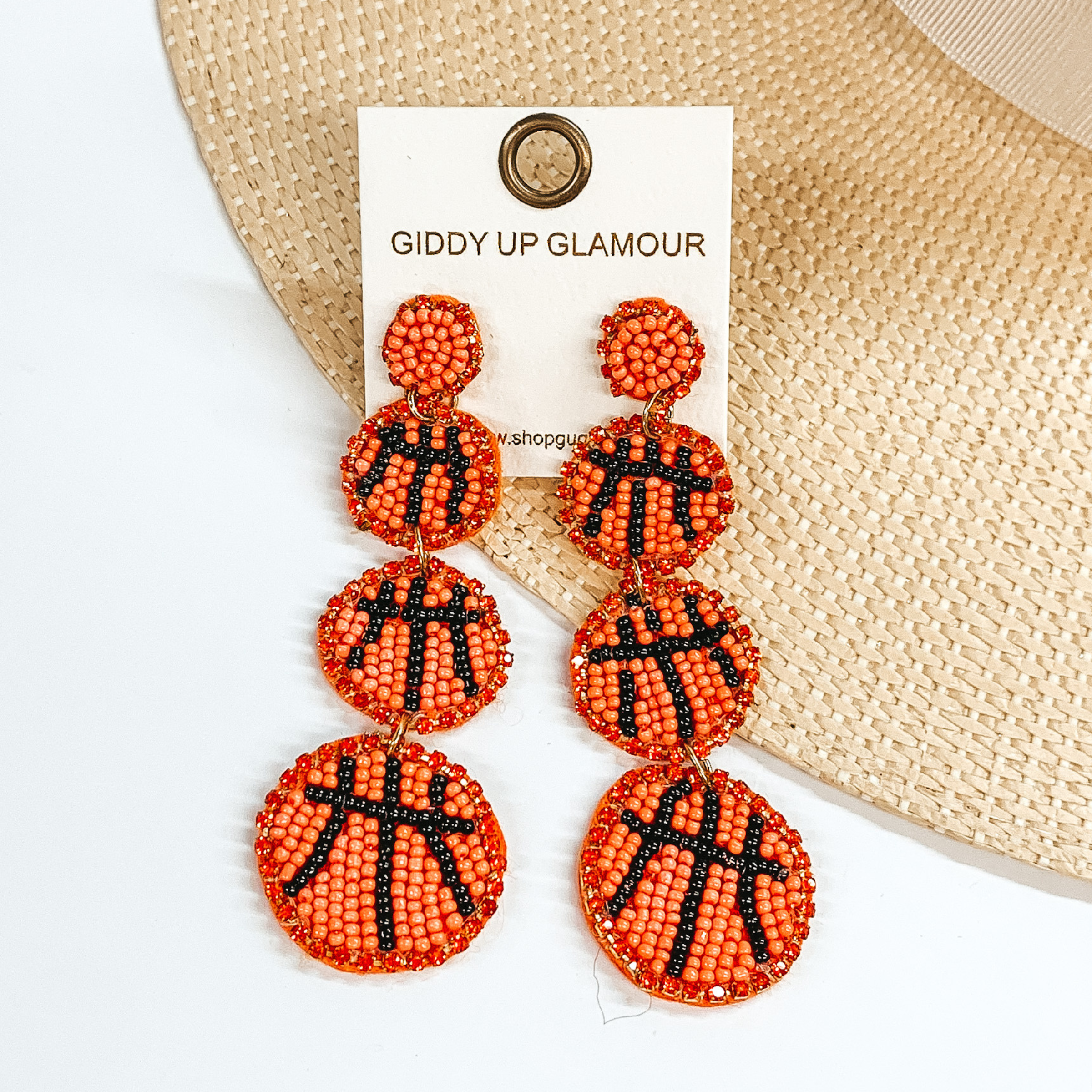 Orange beaded Post back earrings with three beaded basketball pendants. These earrings also include an orange crystal outline. These earrings are pictured partially laying on a straw hat brim on a white background. 