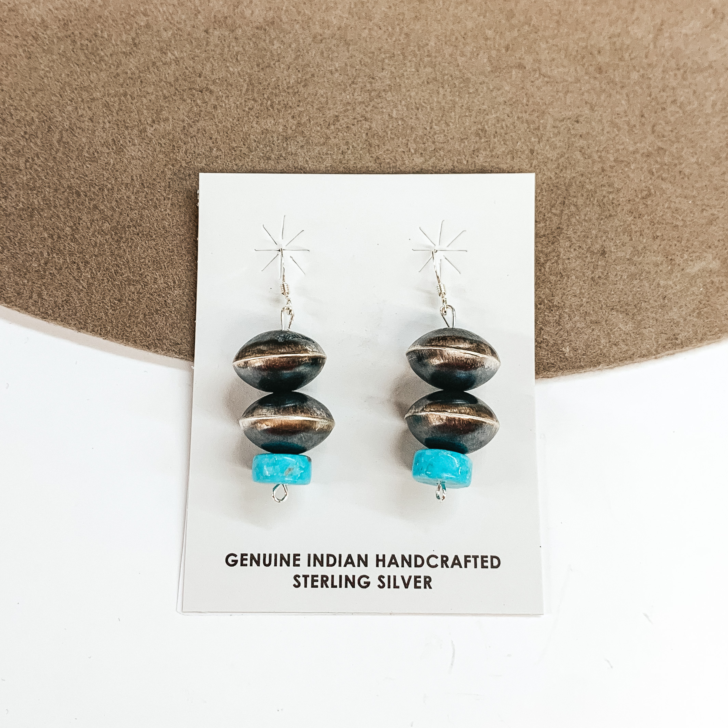The earrings pictured include fish hook earrings with a two saucer pearl drop and a turquoise stone at the bottom. These earrings are pictured on a tan and white background. 