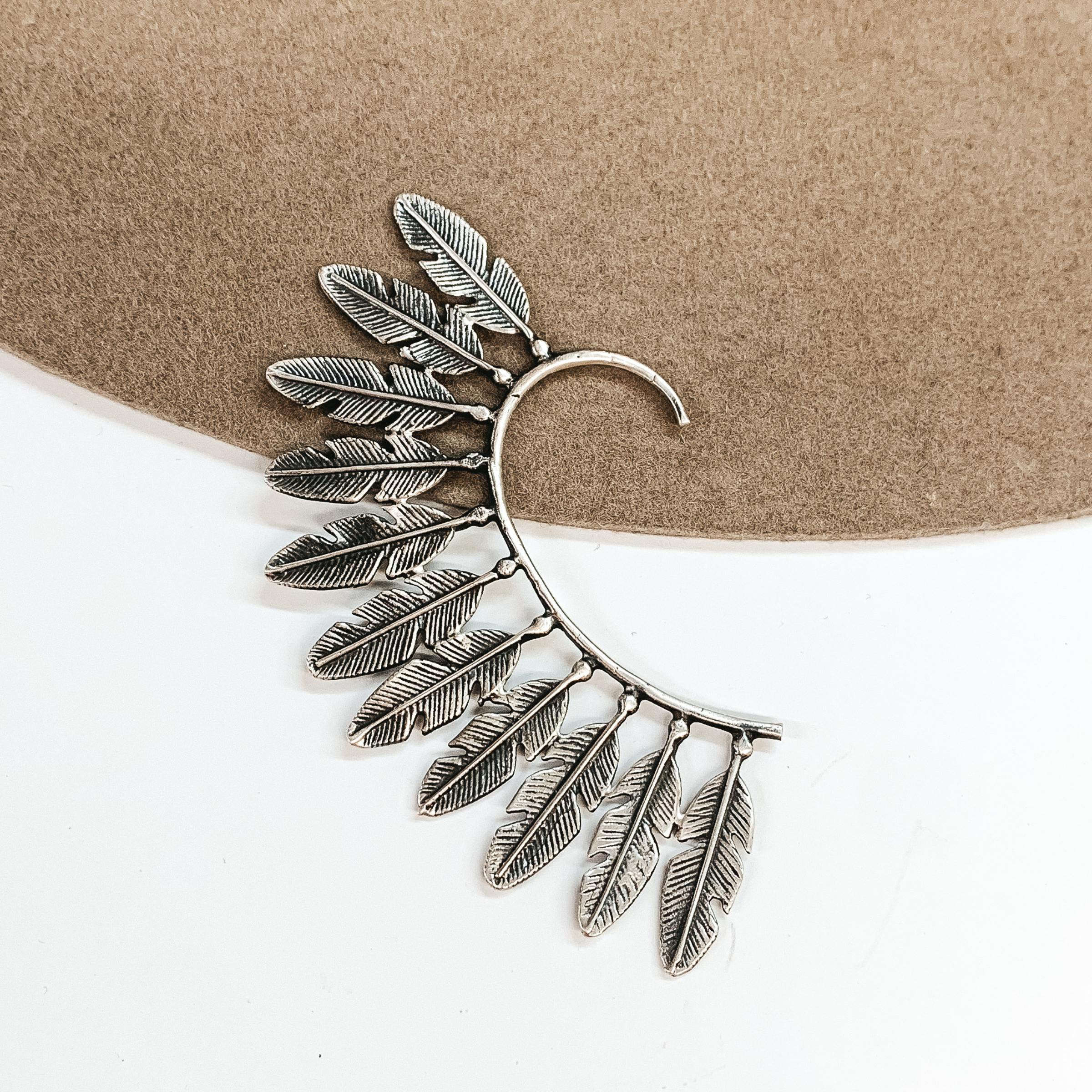 Silver ear cuff with silver feathers outlining the outside of the ear cuff. This earring cuff is pictured on a tan and white background. 