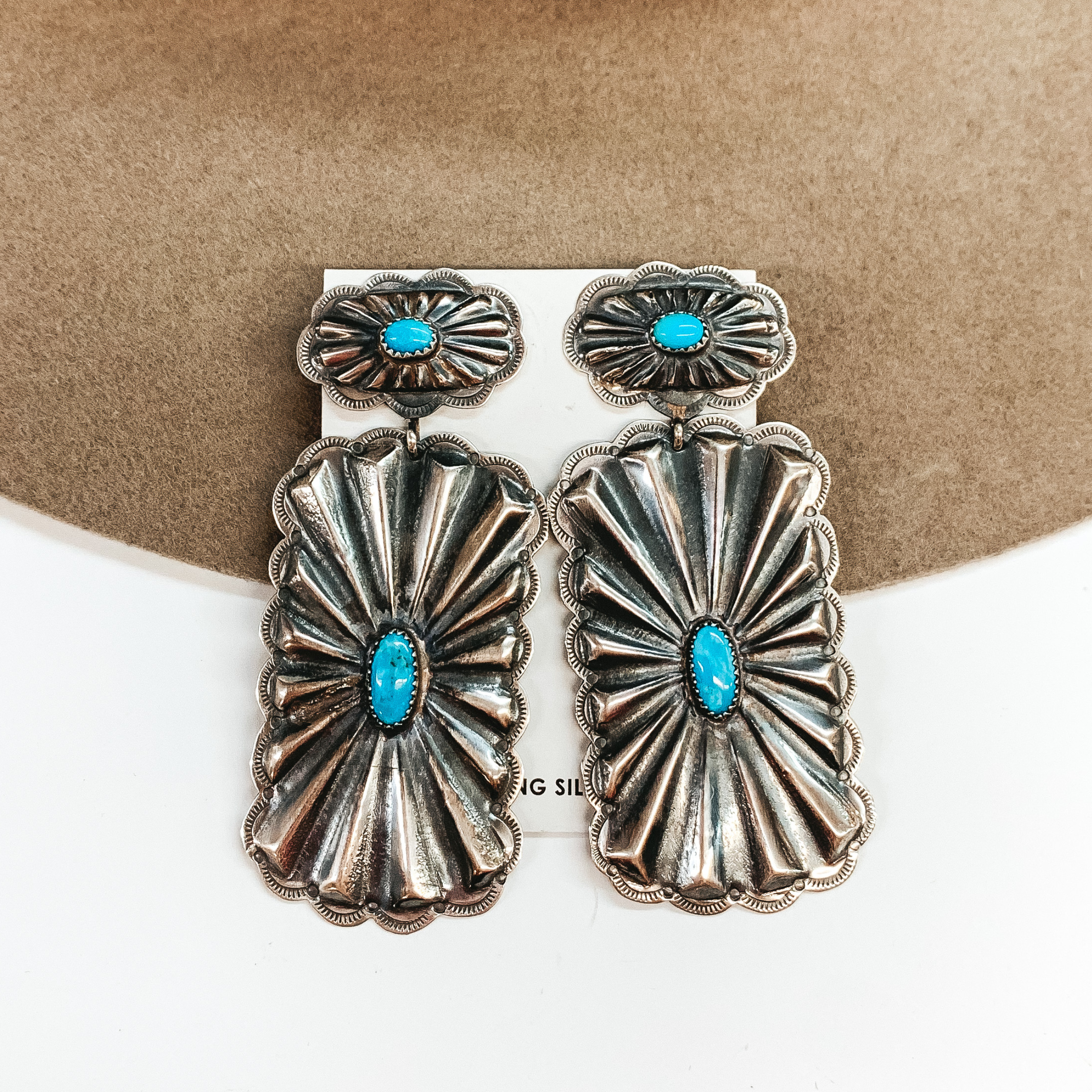 Silver oval concho post earrings with a rectangle concho drop. Both conchos have turquoise stones in the center. These earrings are pictured on a tan and white background. 