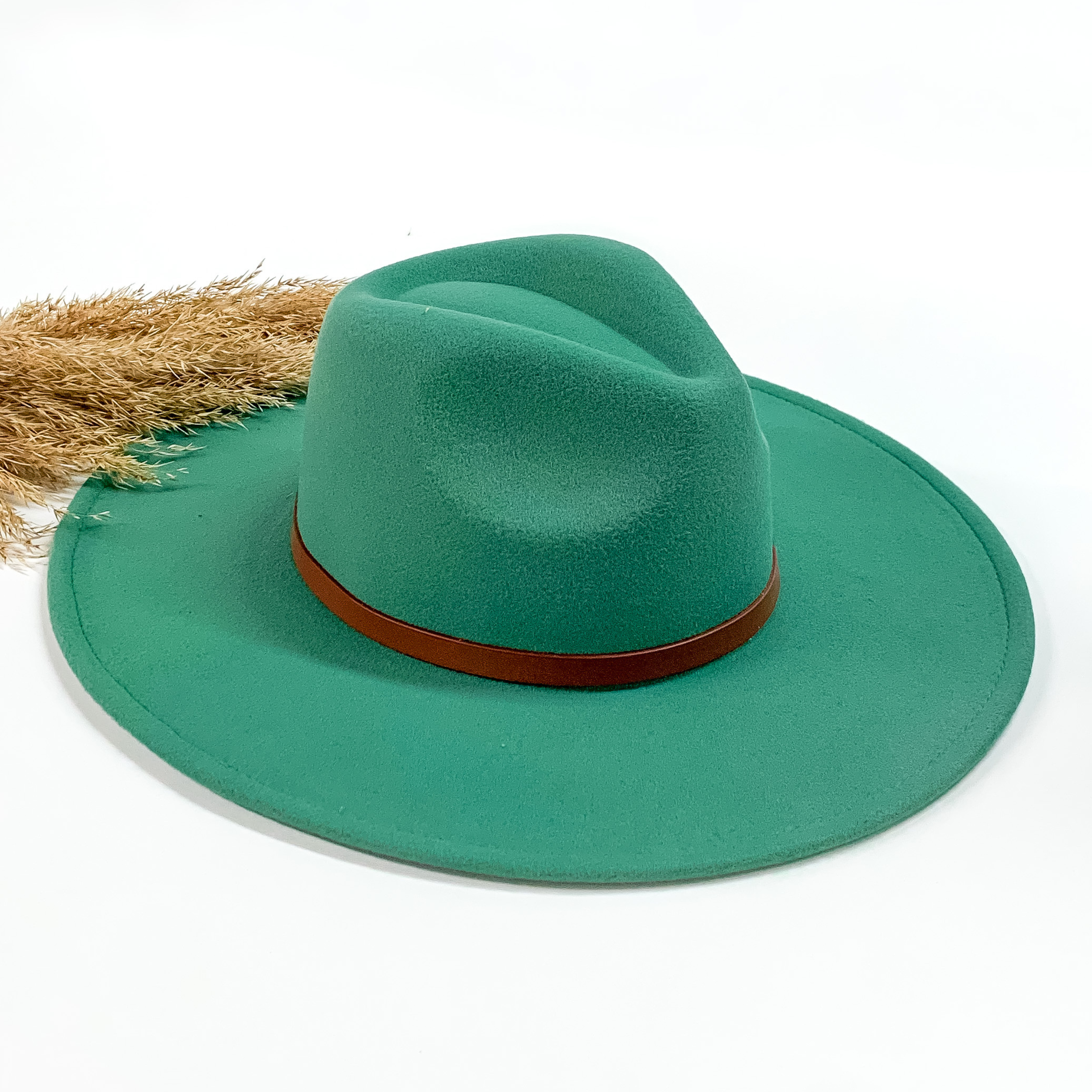 Turquoise faux felt hat with a thin brown hat band. This hat is pictured on a white background with tan pompous in the background.