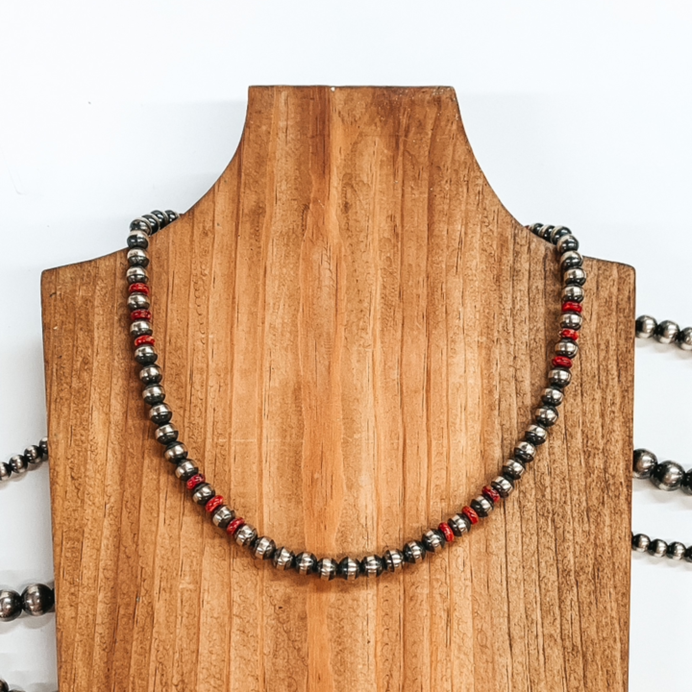 Silver beaded neckace with red bead spacers. This necklace is pictured laying on a wood necklace holder on a white background.